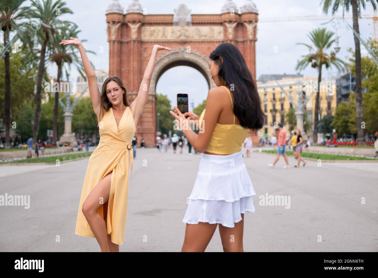 Junge Frau in stilvollem gelben Kleid, die mit den Armen vor dem historischen Triumphbogen aufliegt, während asiatische Freundin am Sommertag während der Stadtbesichtigung in Barcelona auf dem Smartphone fotografiert Stockfoto