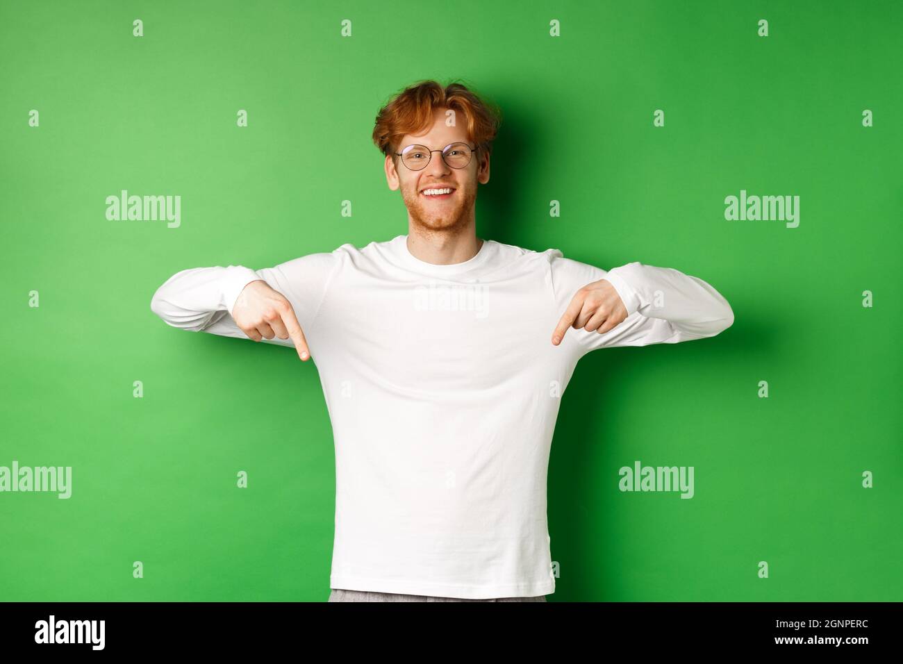 Gutaussehendes männliches Modell mit roten Haaren, Brille und Langarm tragend, Finger nach unten zeigend und fröhlich lächelnd, zeigt promo, grüner Hintergrund Stockfoto