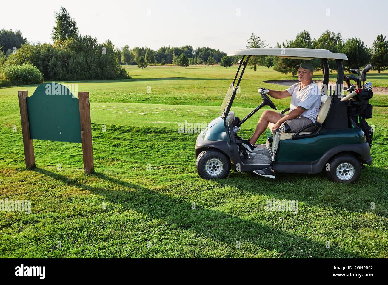 Golfer mit Beinprothese, der an einem sonnigen Tag auf dem Rasen auf dem Golfplatz in der Nähe des Namensschilds des Golfclubs über Landschaft und Fairway Golf Cart fährt Stockfoto