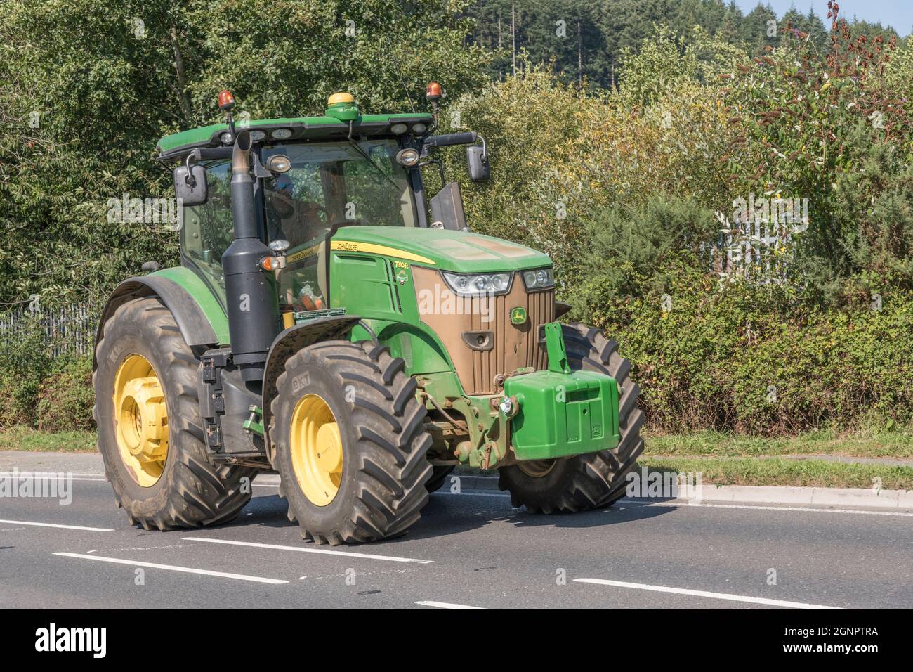 Bekannter grüner John Deere-Traktor auf offener Landstraße, die bergauf führt. Großreifen-Modell von Deere ist 7310 R. Stockfoto