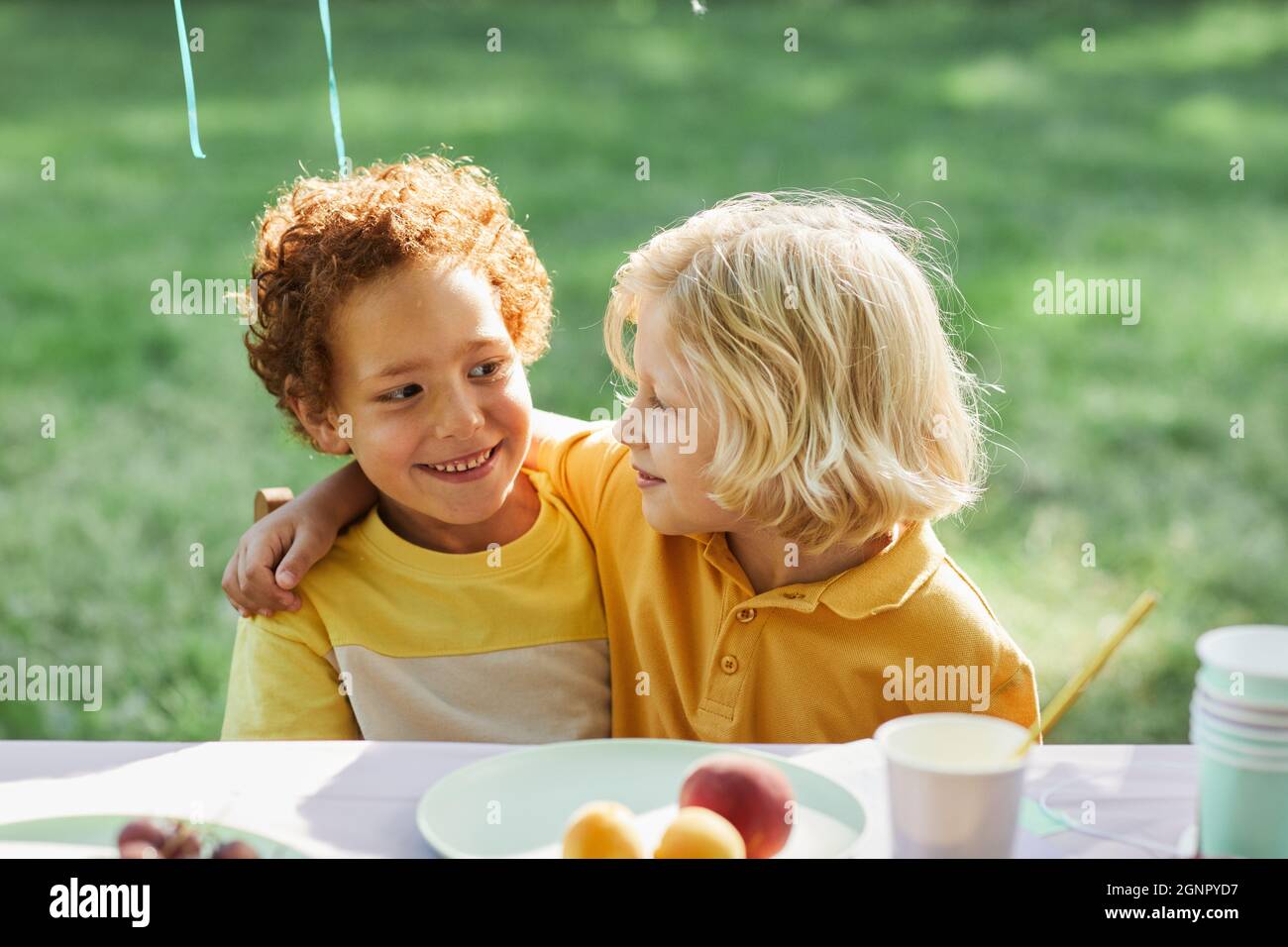 Portrait von zwei lächelnden Jungen, die sich am Picknicktisch im Freien umarmen, während sie die Geburtstagsfeier im Sommer genießen und Platz kopieren Stockfoto
