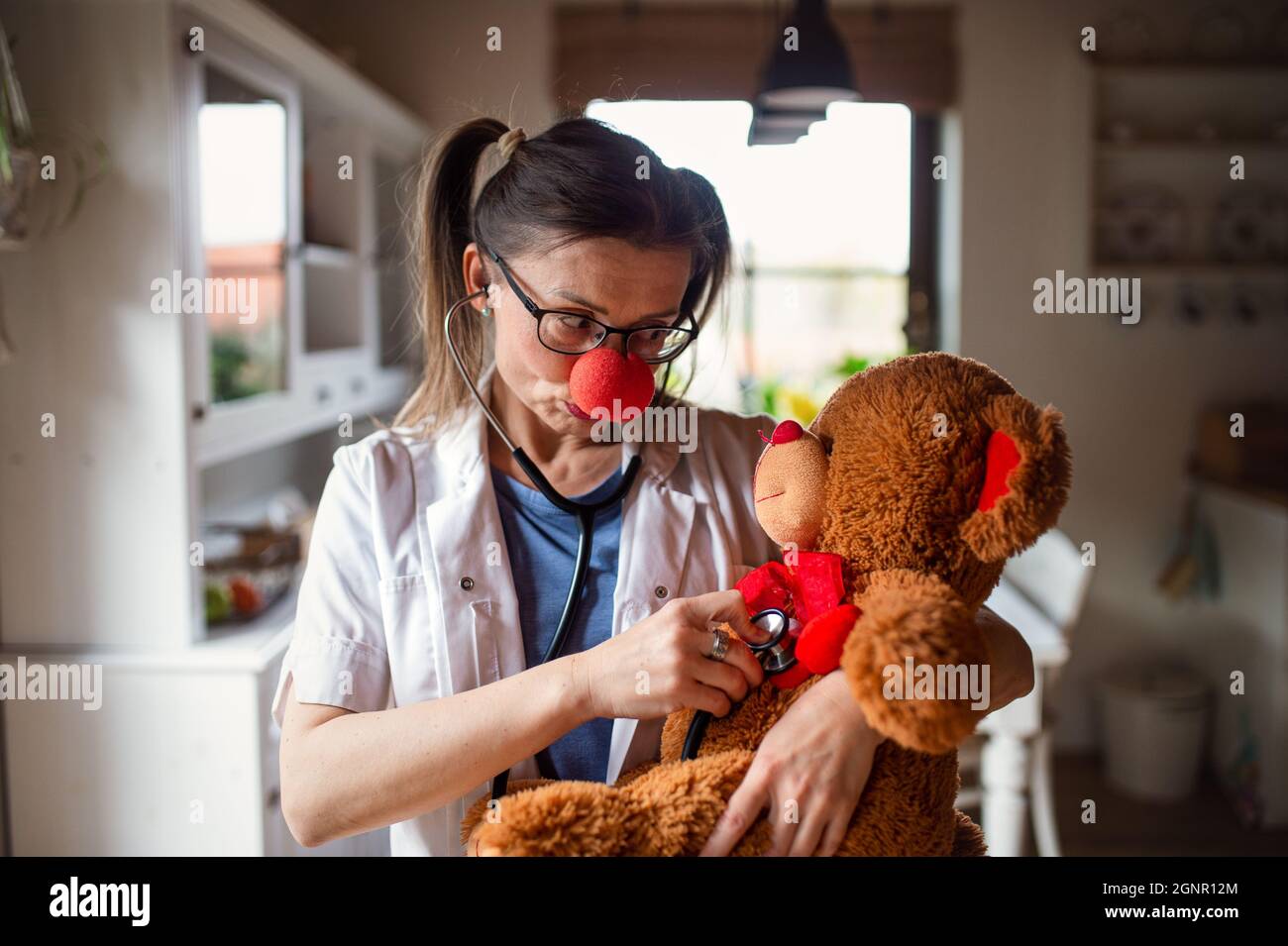 Porträt einer lustigen Ärztin mit roter Clownnase, die einen Teddybären hält. Stockfoto