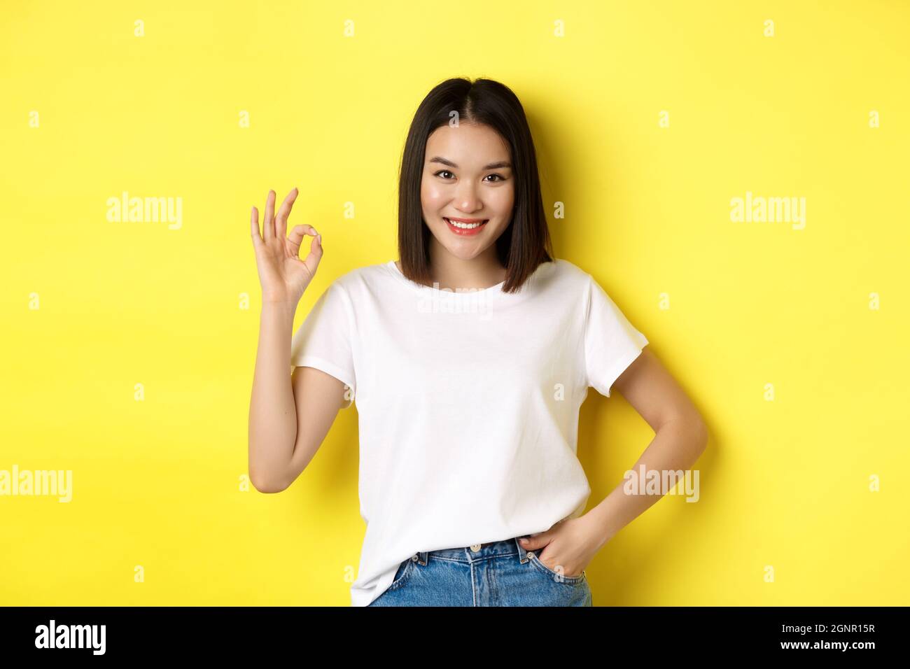 Zuversichtlich asiatische Frau lächelnd und zeigt OK Zeichen, genehmigen und loben gutes Angebot, stehend in weißem T-Shirt über gelbem Hintergrund Stockfoto