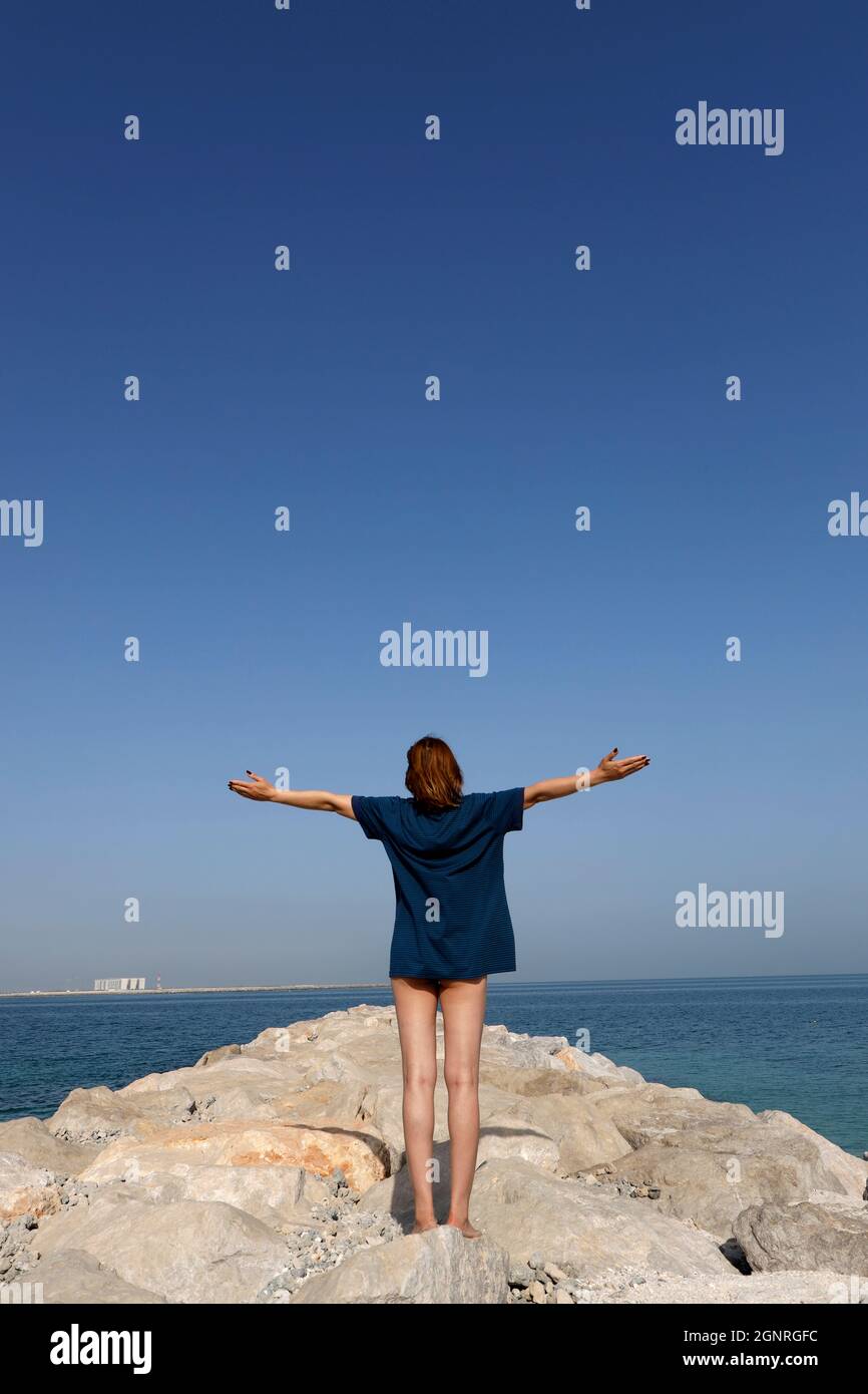 Frau im weißen T-Shirt, die ihre Arme mit Blick auf das Meer hebt. Vereinigte Arabische Emirate. Stockfoto