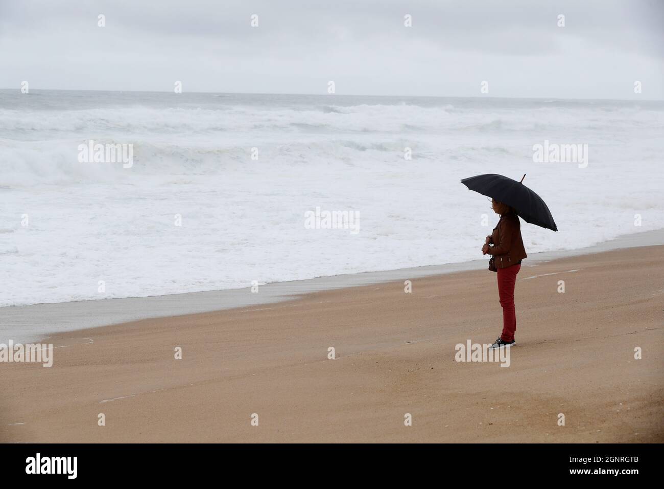 Frau mit schwarzem Regenschirm, die an einem launischen und regnerischen Tag vor der Küste steht. Nazare. Portugal. Stockfoto