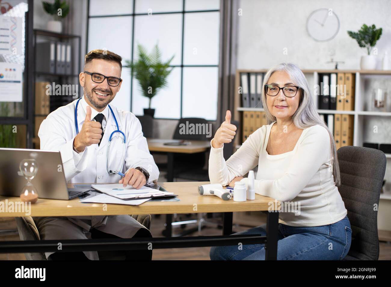 Kaukasischer bärtiger Arzt in weißem Laborkittel und Brille sitzt mit einer älteren Frau am Schreibtisch, lächelt vor der Kamera und zeigt den Daumen hoch. Konzept von Menschen, Behandlung und Gesundheitsfürsorge. Stockfoto