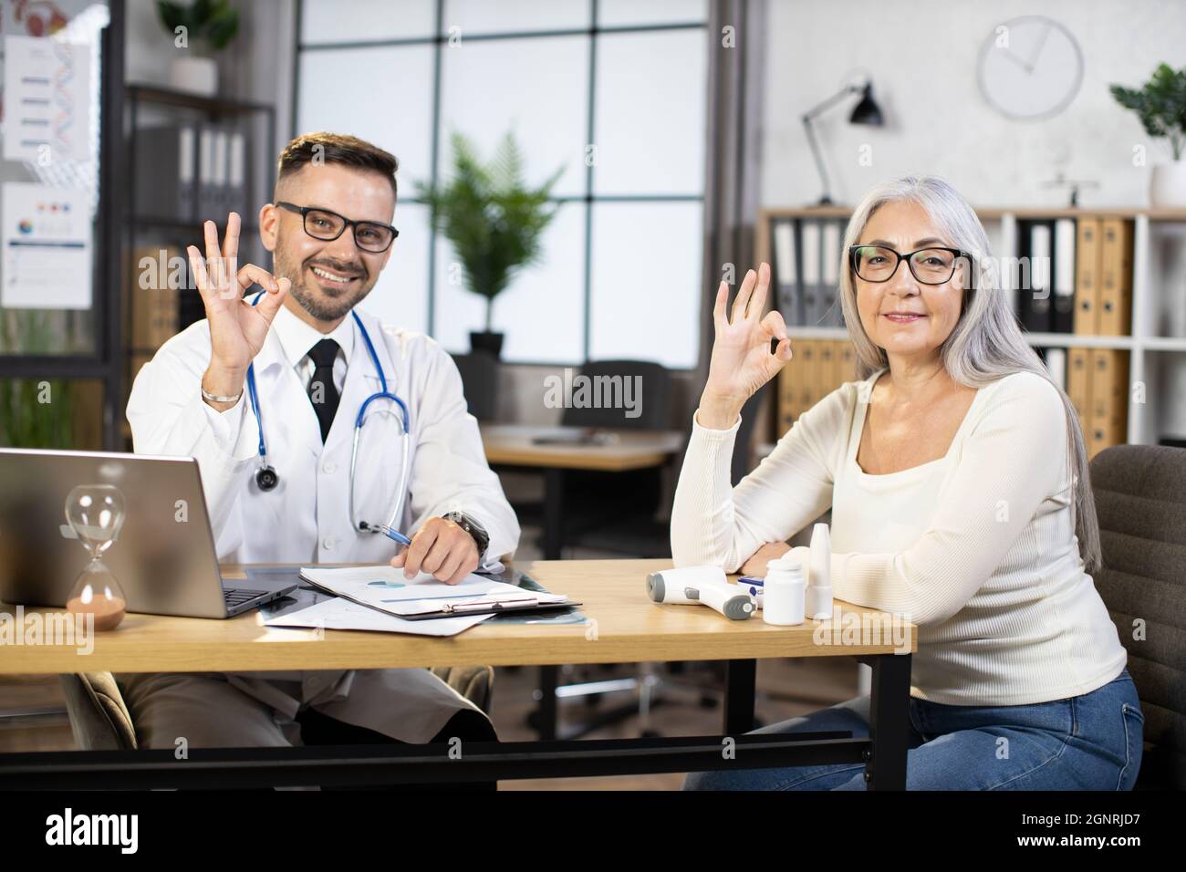 Kaukasischer bärtiger Arzt in weißem Laborkittel und Brillen, der mit einer pensionierten Dame am Schreibtisch saß, vor der Kamera lächelte und ein OK cooles Schild zeigte. Konzept von Menschen, Behandlung und Gesundheitsfürsorge. Stockfoto