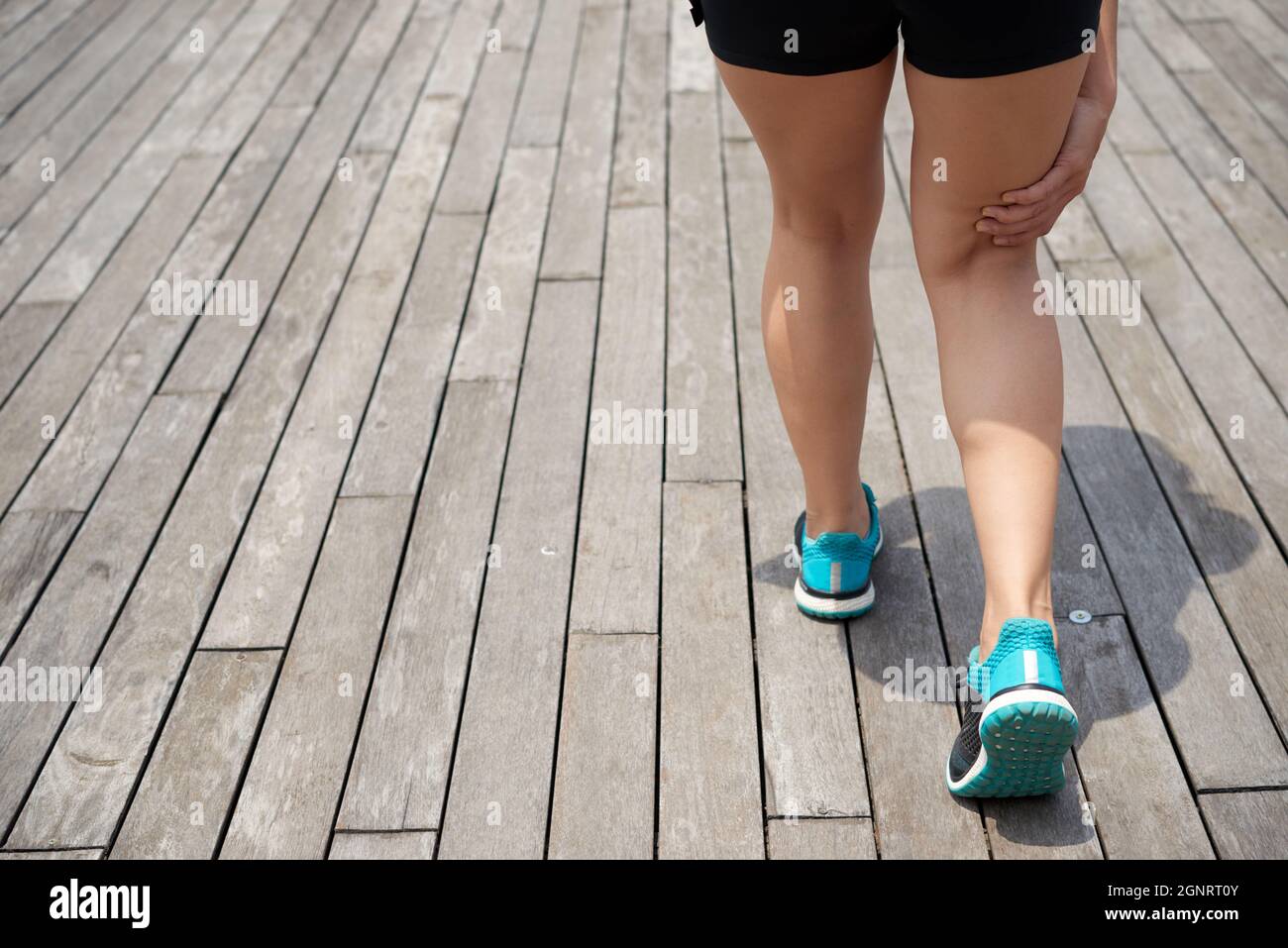 Rückansicht einer unerkennbaren Läuferin in blauen Sportschuhen, die auf einem hölzernen Pier steht und den Schmerzpunkt berührt, während sie unter der Kniescheibe Schmerzen spürt Stockfoto
