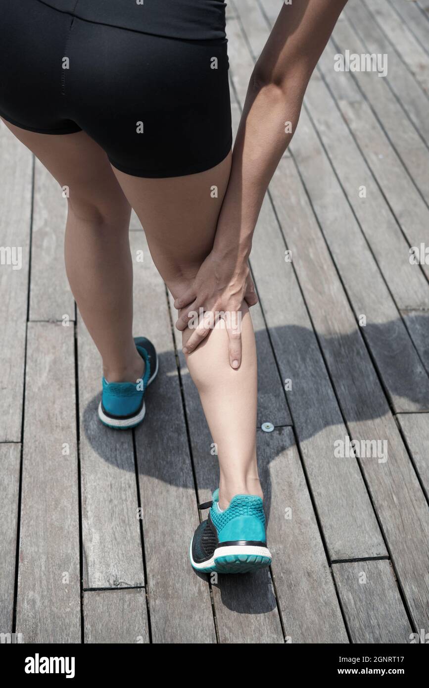 Nahaufnahme einer nicht erkennbaren Frau in kurzen Hosen und einem blauen Sportschuh, die schmerzhaftes Bein berührt, während sie auf dem hölzernen Pier läuft Stockfoto