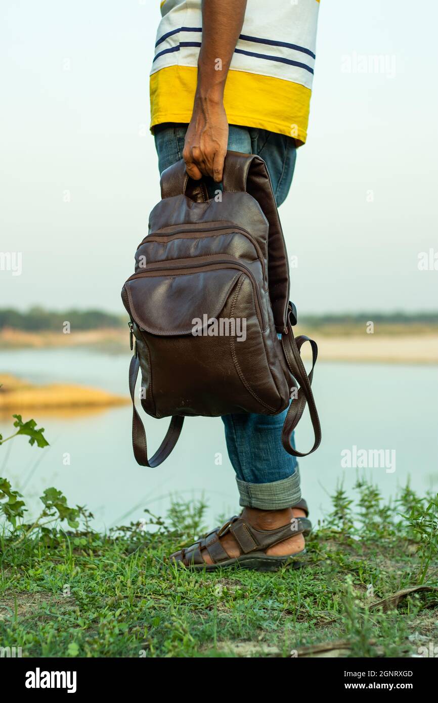 Der Mann mit einem Laptop, der eine multifunktionale Ledertasche trägt oder reist und allein am Ufer des Flusses steht Stockfoto