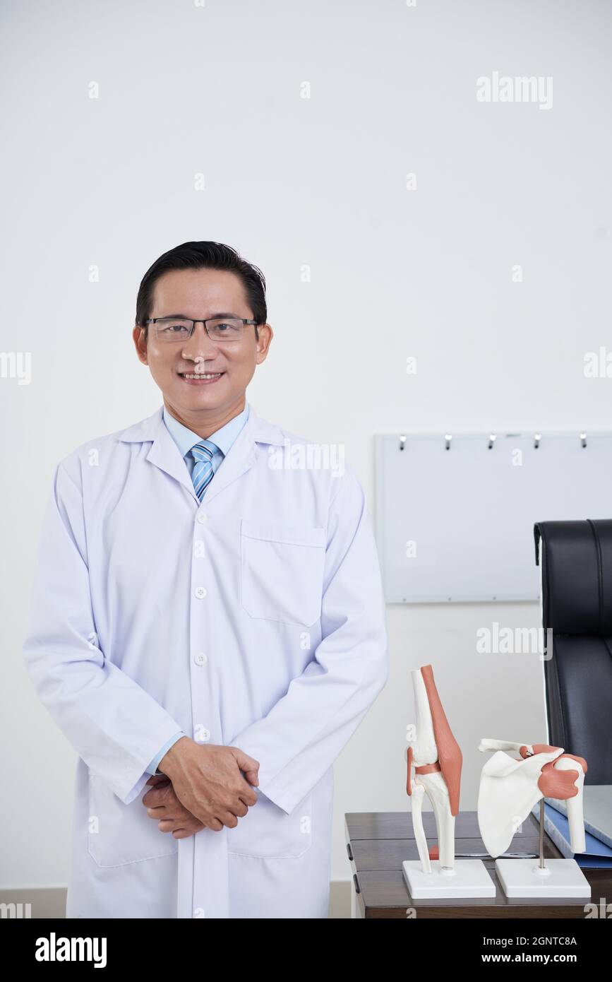 Vertikales mittleres Porträt eines reifen asiatischen Arztes mit weißem Mantel und Brille, der in seinem Büro mit einem Lächeln auf die Kamera steht Stockfoto