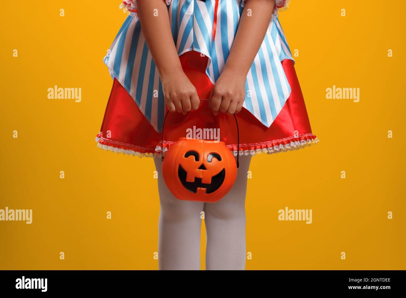 Kleines Mädchen in einem Faschingskostüm für Halloween auf gelbem Hintergrund. Das Kind hält einen Eimer mit einer Kürbislaterne. Stockfoto