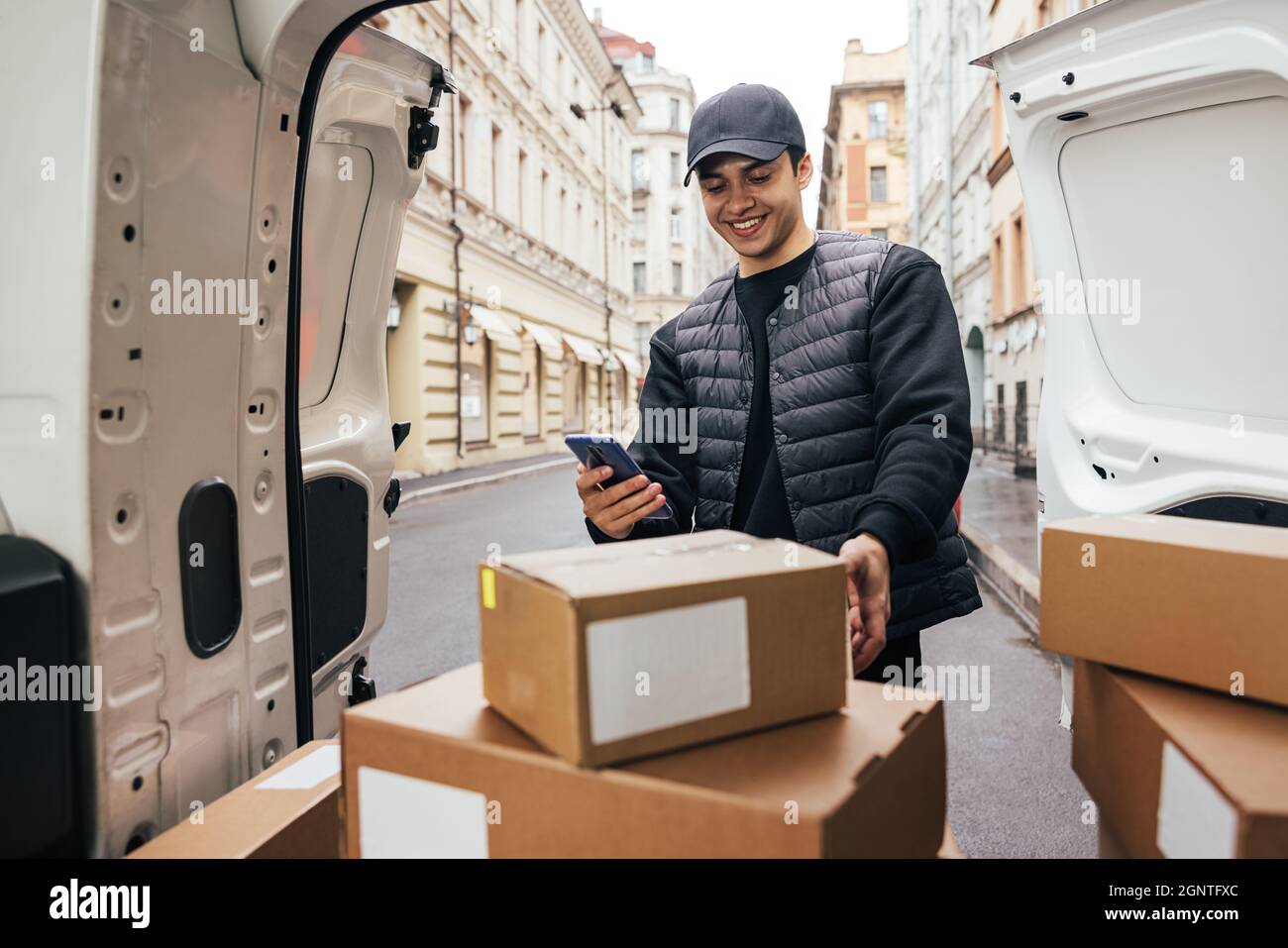 Lächelnder Kurier in Uniform, der am Kofferraum des Autos stand und das Mobiltelefon auf Kartons blickte Stockfoto