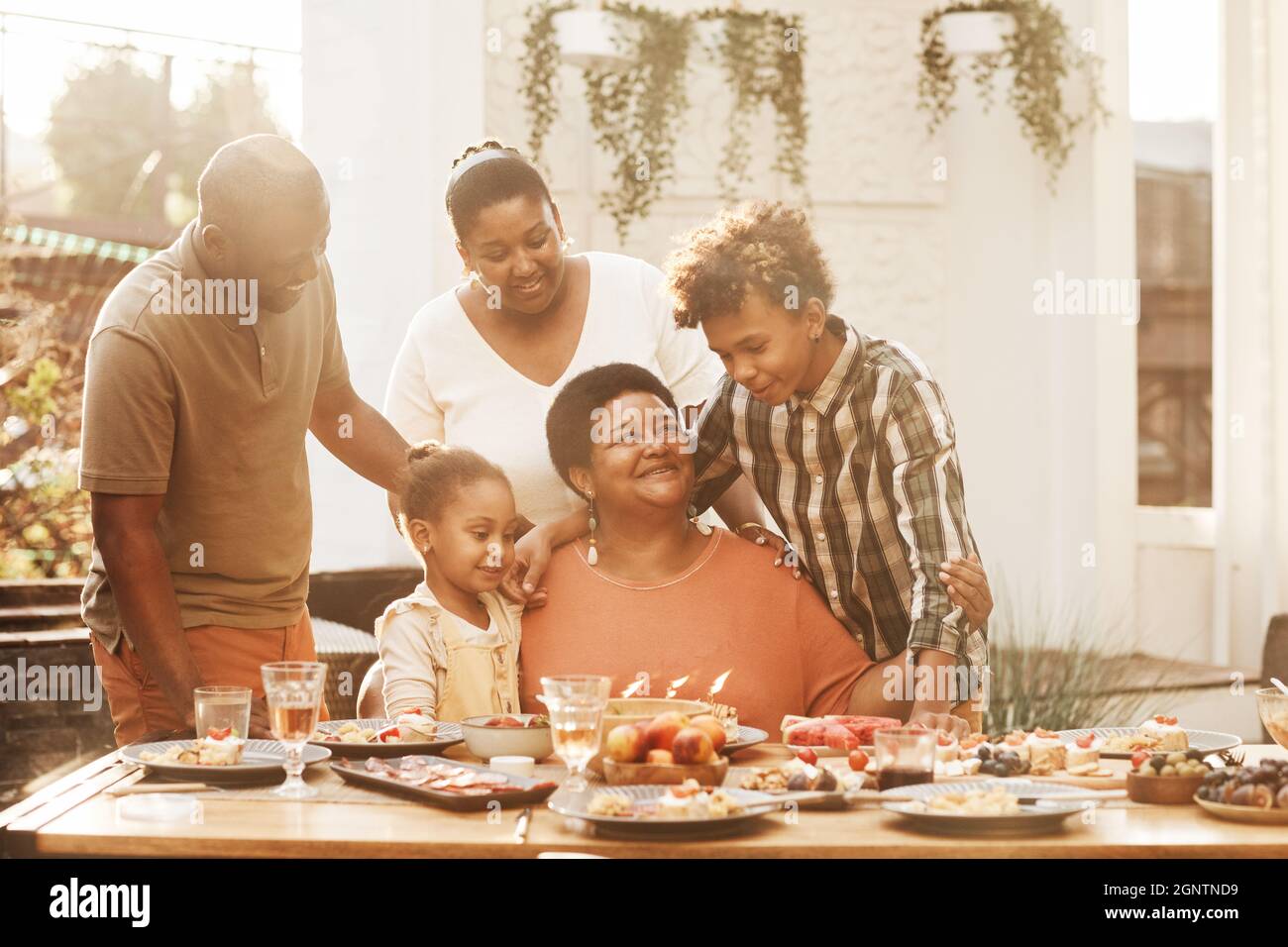 Porträt einer glücklichen afroamerikanischen Großmutter, die mit der Familie beim Abendessen im Freien Geburtstag feiert, beleuchtet von Sonnenlicht Stockfoto