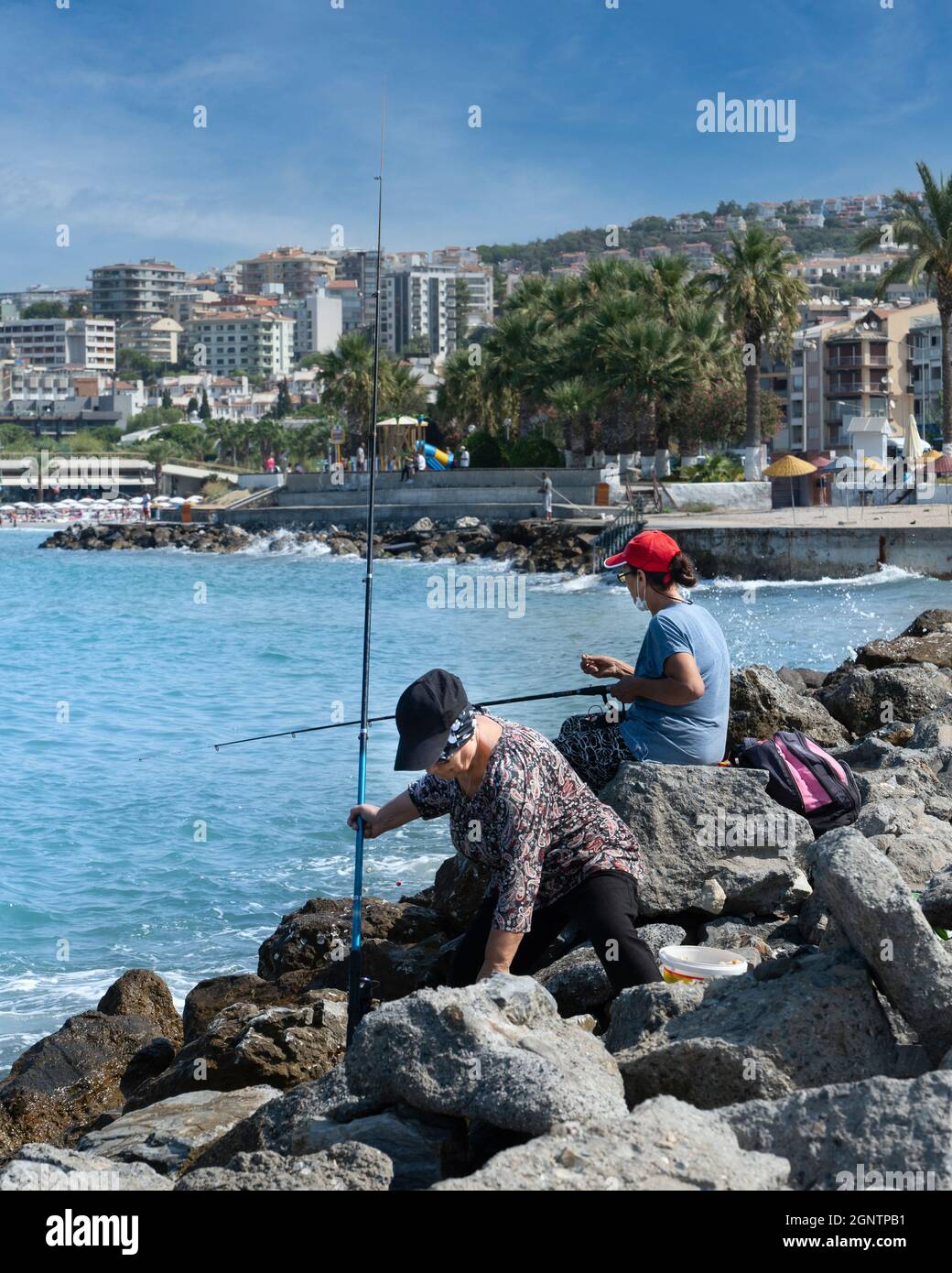 Kusadasi, Aydin, Türkei - 23. August 2021: Zwei Frauen sitzen auf Felsen am Meer und fischen. Stockfoto
