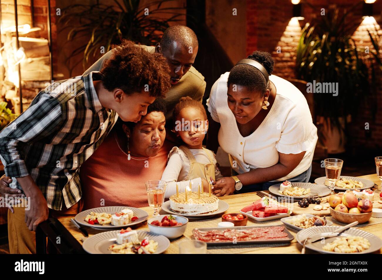 Porträt einer afroamerikanischen Oma, die Kerzen bläst, während sie mit der Familie in gemütlicher Atmosphäre Geburtstag feiert Stockfoto