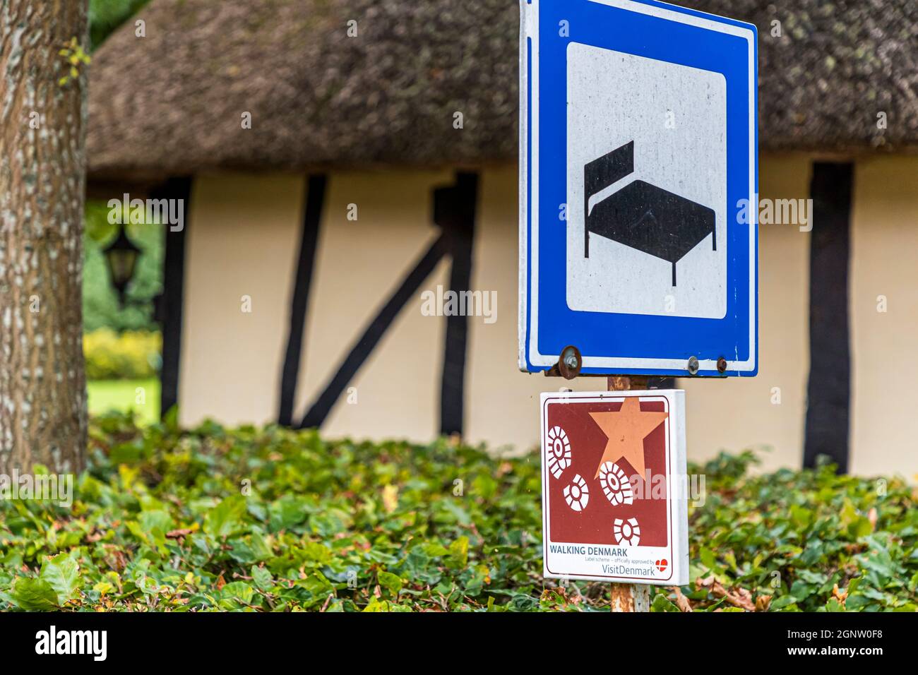 Ein blaues Schild mit einem Bettsymbol markiert eine Fachwerkhausunterkunft entlang der Øhavsstien nahe Svendborg, Dänemark; ein Aufkleber der Kampagne „Walking Denmark“ ist darunter angebracht. Stockfoto