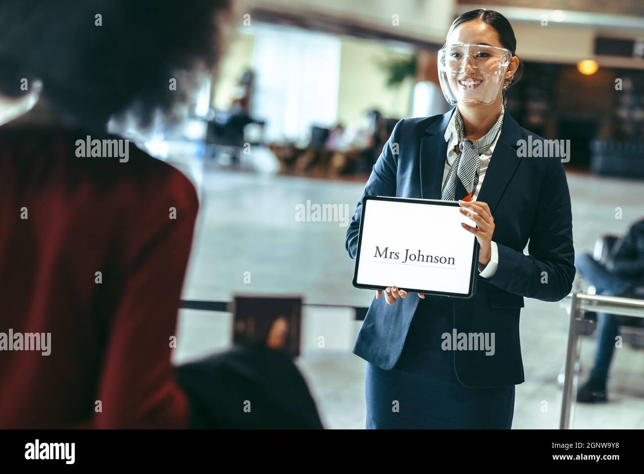 Eine Fahrerin mit Gesichtsschutz, die am Flughafenterminal mit einem  Namensschild steht, um einen Reisenden zu empfangen. Chauffeur mit einem  Schild am Ankunftsterminal Stockfotografie - Alamy