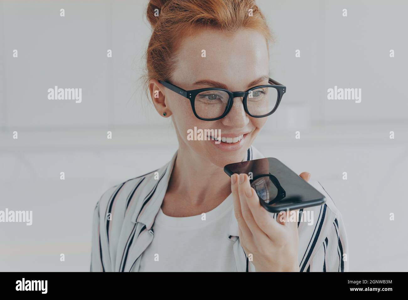 Rotschopf Frau in durchsichtiger Brille spricht mit virtuellen Assistenten hält Handy in der Nähe des Mundes Stockfoto