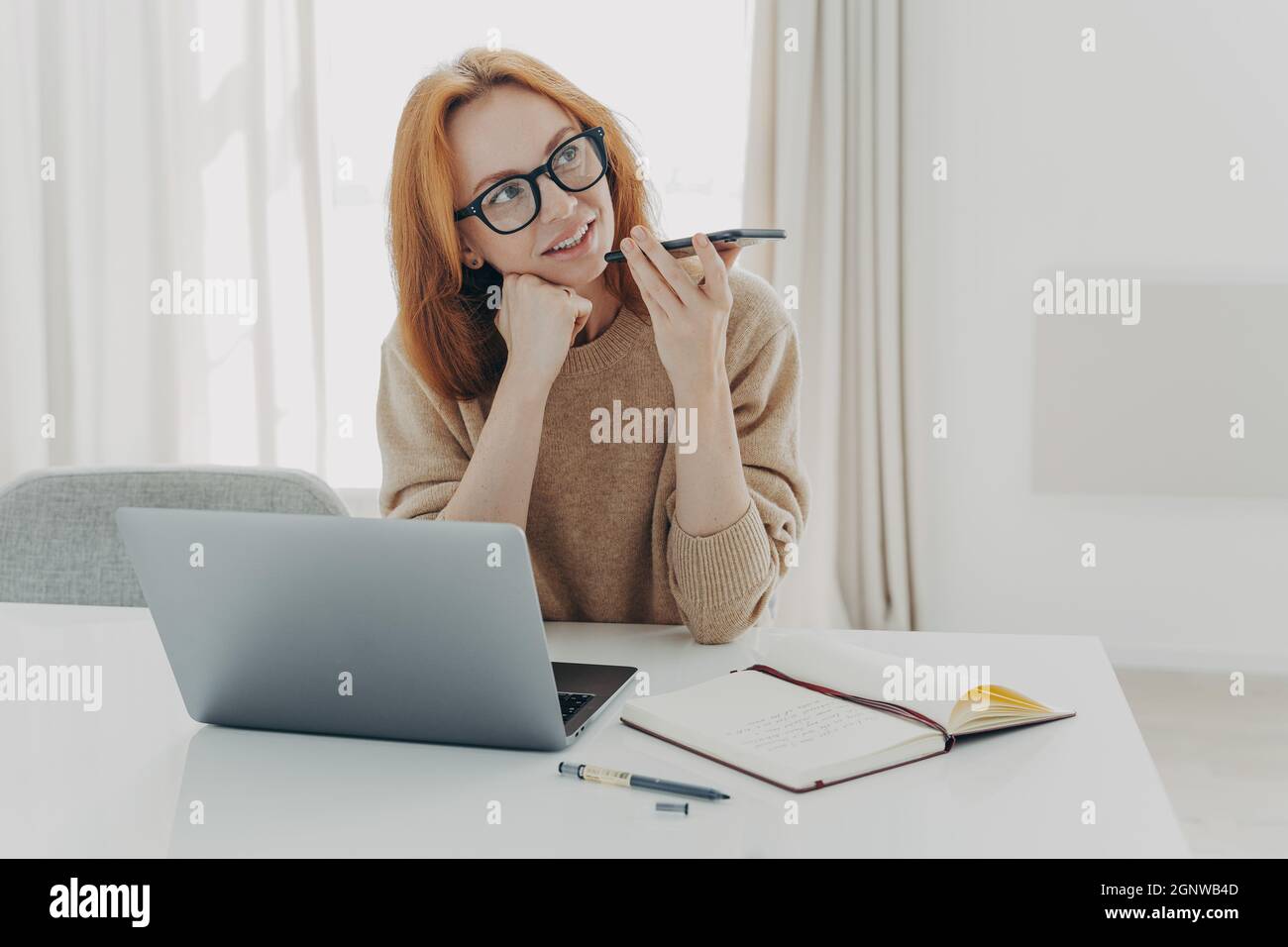 Freiberufliche Mitarbeiterin nimmt Sprachnachrichten auf, die auf dem Schreibtisch sitzen und Notizen auf dem Notizblock machen Stockfoto