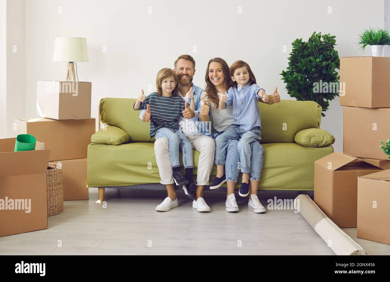 Glückliche Familie mit Kindern, die in ihrem neuen Zuhause auf der Couch sitzen und Daumen nach oben geben Stockfoto