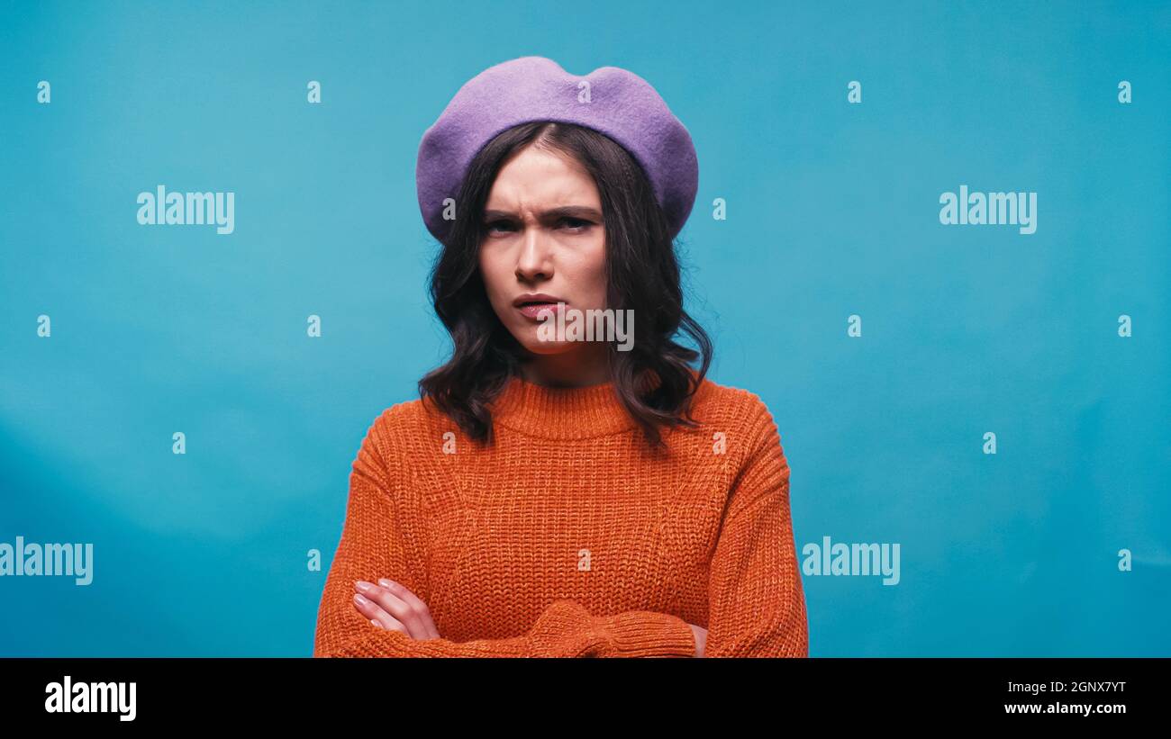 Misstrauische Frau in violettem Baskenmütze und orangefarbenem Pullover, die die Kamera isoliert auf Blau anschaut Stockfoto