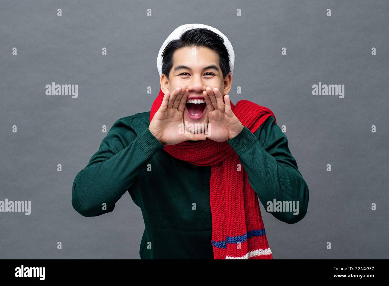 Fröhlicher junger asiatischer Mann in weihnachtlicher Kleidung, schreiend mit Händen, die um den Mund in grauem, isoliertem Studiohintergrund schröpfen Stockfoto