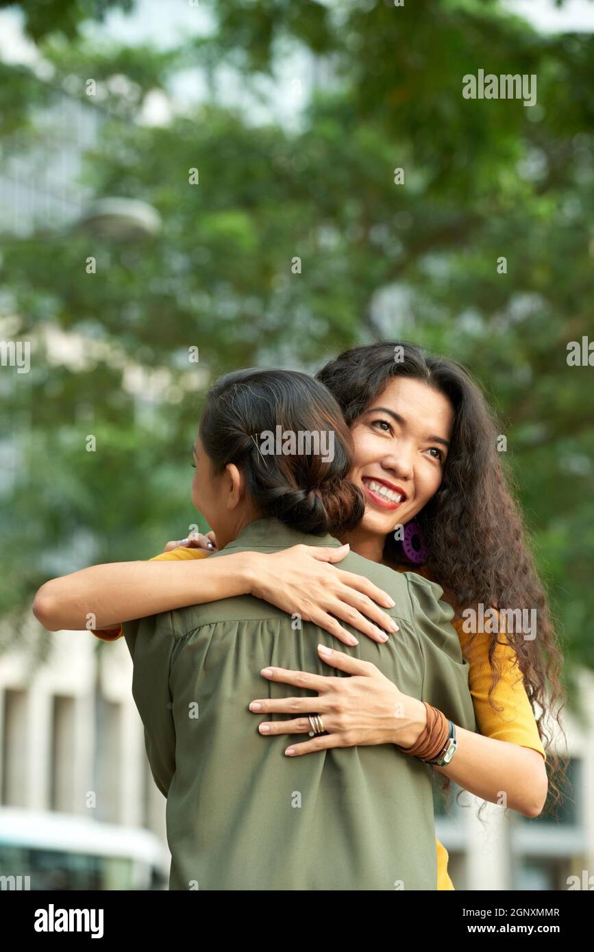 Lächelnde Erwachsene asiatische Tochter mit lockigen Haaren umarmte Mutter im Sommerpark, während sie sich mit ihr trifft Stockfoto