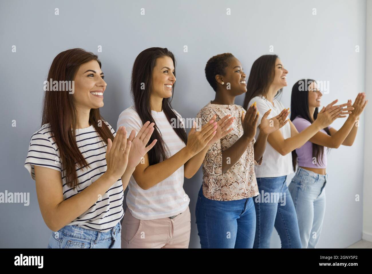 Eine Gruppe glücklicher junger multiethnischer Frauen, die an der Wand stehen, lächeln und applaudieren Stockfoto
