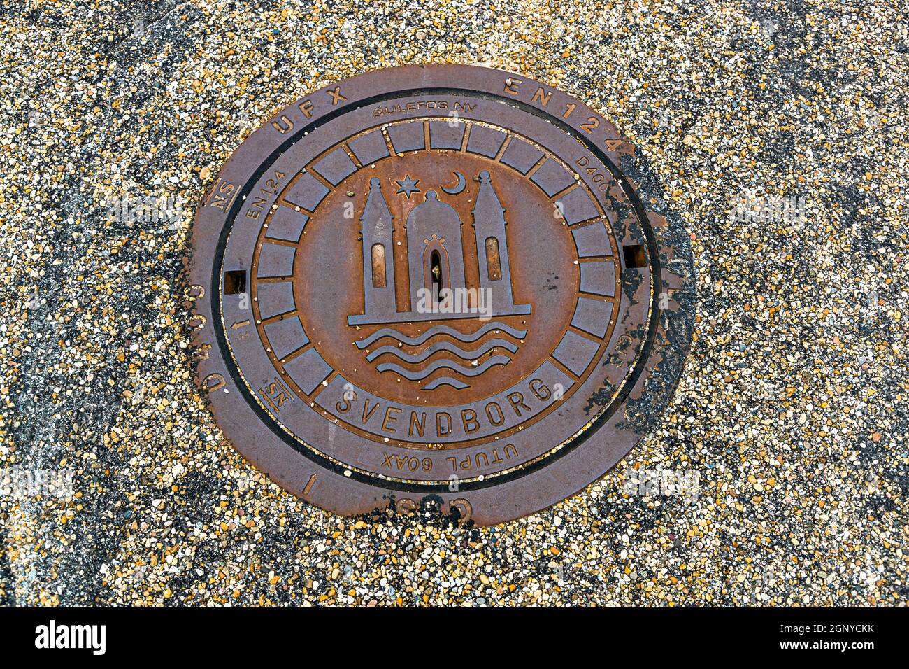 Ein Gullydeckel in Svendborg, Dänemark, trägt das Wappen der Stadt: drei Türme über welligen Linien, mit einem Stern und einem Halbmond darüber. Stockfoto
