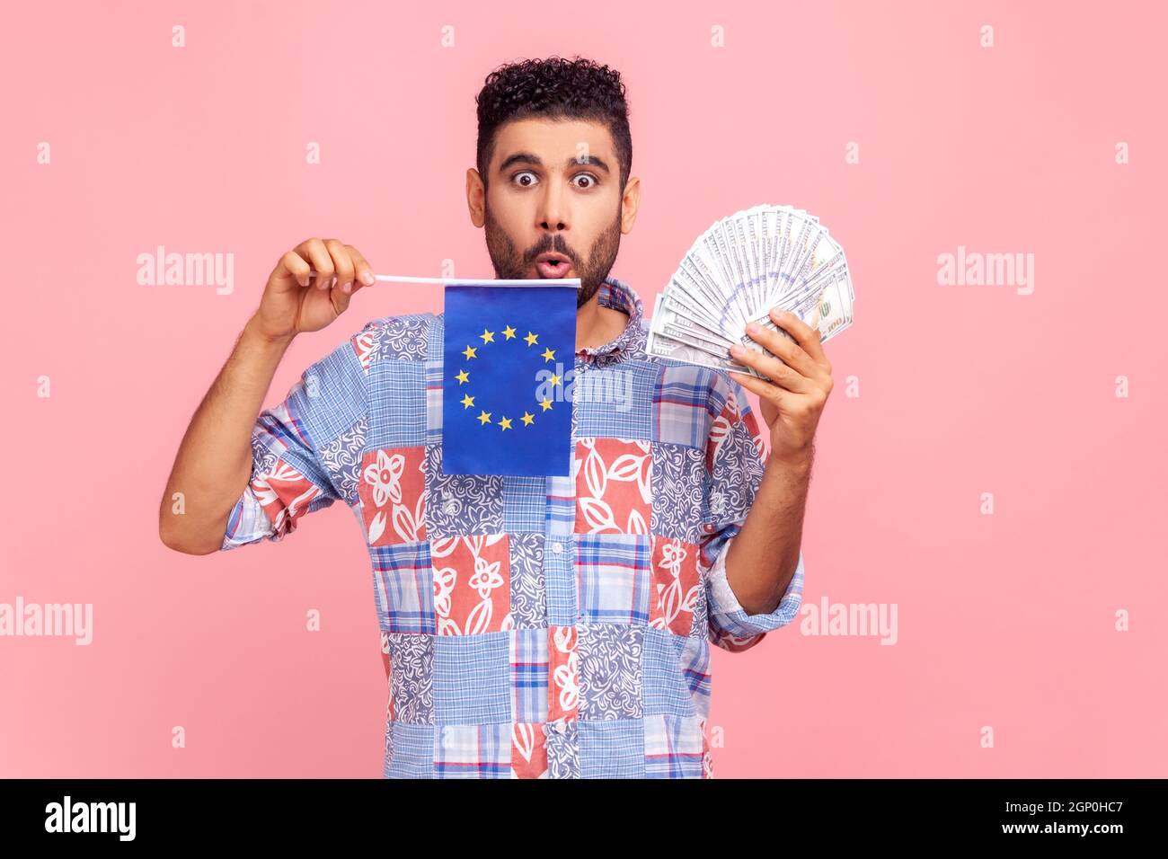 Porträt eines staunenden bärtigen Mannes, der ein blaues Hemd im Casual-Stil trägt, eine Kamera mit großen schockierten Augen sieht und die europäische Flagge und die Banknoten hält. Innenaufnahme des Studios isoliert auf rosa Hintergrund. Stockfoto