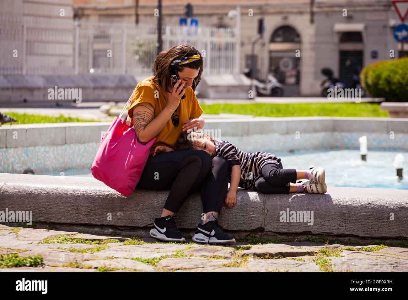 Triest, Italien - Mai 31: Mutter und kleine Tochter sitzen auf der Bank. Mama telefoniert, während sie ihre kleine Tochter auf ihrem Schoß kuschelt Stockfoto