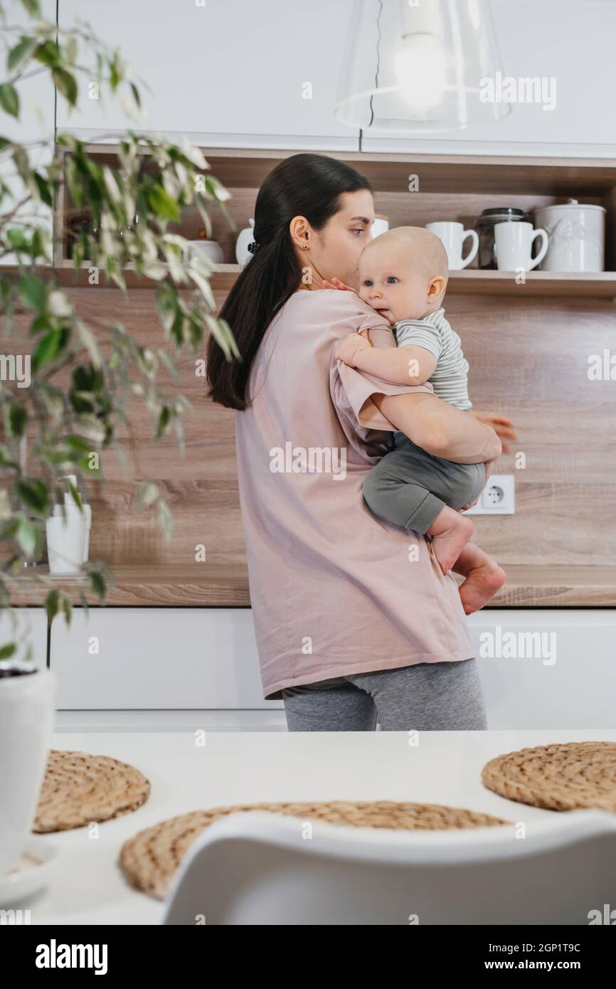 Beschäftigt Mutter Reinigung Küche mit Baby auf den Armen. Frau kocht und hält Säugling, müde Frau macht tägliche Hausaufgaben mit Kind. Stockfoto