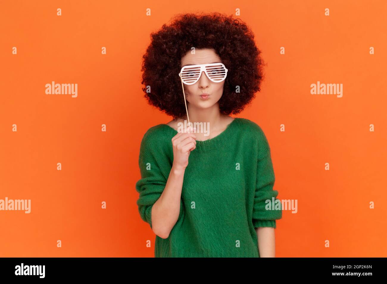Verspielte Frau mit Afro-Frisur trägt grünen legeren Stil Pullover stehend Brille auf Stick, festliche Stimmung, Party, Feier. Innenaufnahme des Studios isoliert auf orangefarbenem Hintergrund. Stockfoto