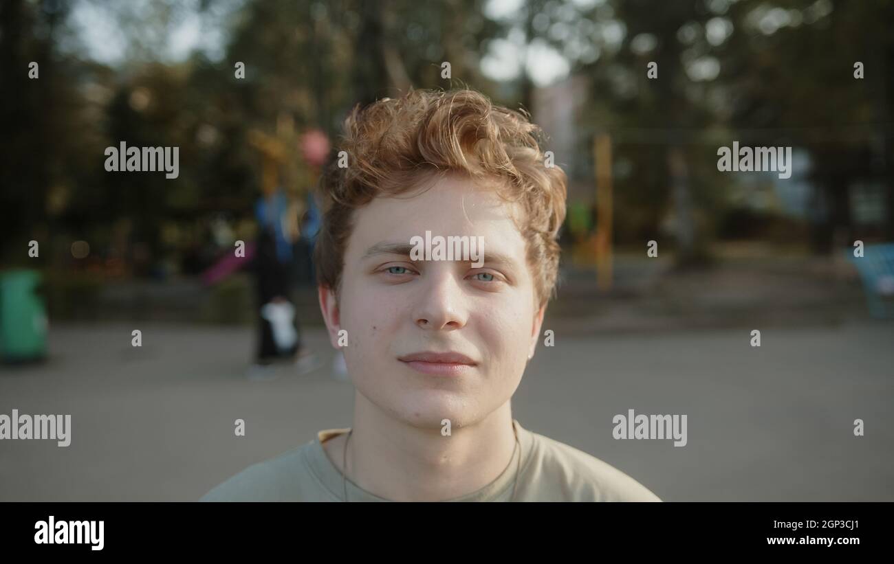 Junger hübscher, blonder, haariger, mit Augen gefärbter Junge, der im Park steht, die Kamera anschaut und lächelt Stockfoto