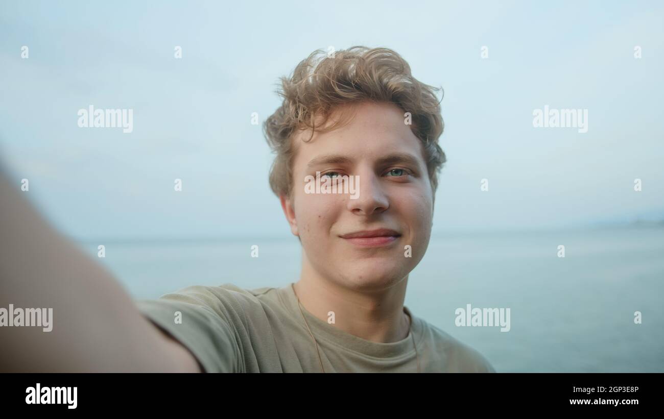 Junge, hübsche blonde, haarfarbene Augen, ein Mann, der mit dem Telefon in der Hand einen Videoanruf abruft oder vloggt, winkt der Kamera zu und lächelt. Meer zurück Stockfoto