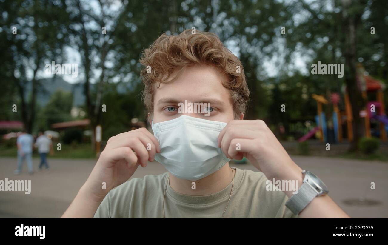 Der blonde junge, gutaussehende Mann zieht die medizinische Maske aus und blickt lächelnd auf die Kamera. Outdoor-Park im Hintergrund. Gesundheitsfürsorge und medizinisches Konzept Stockfoto