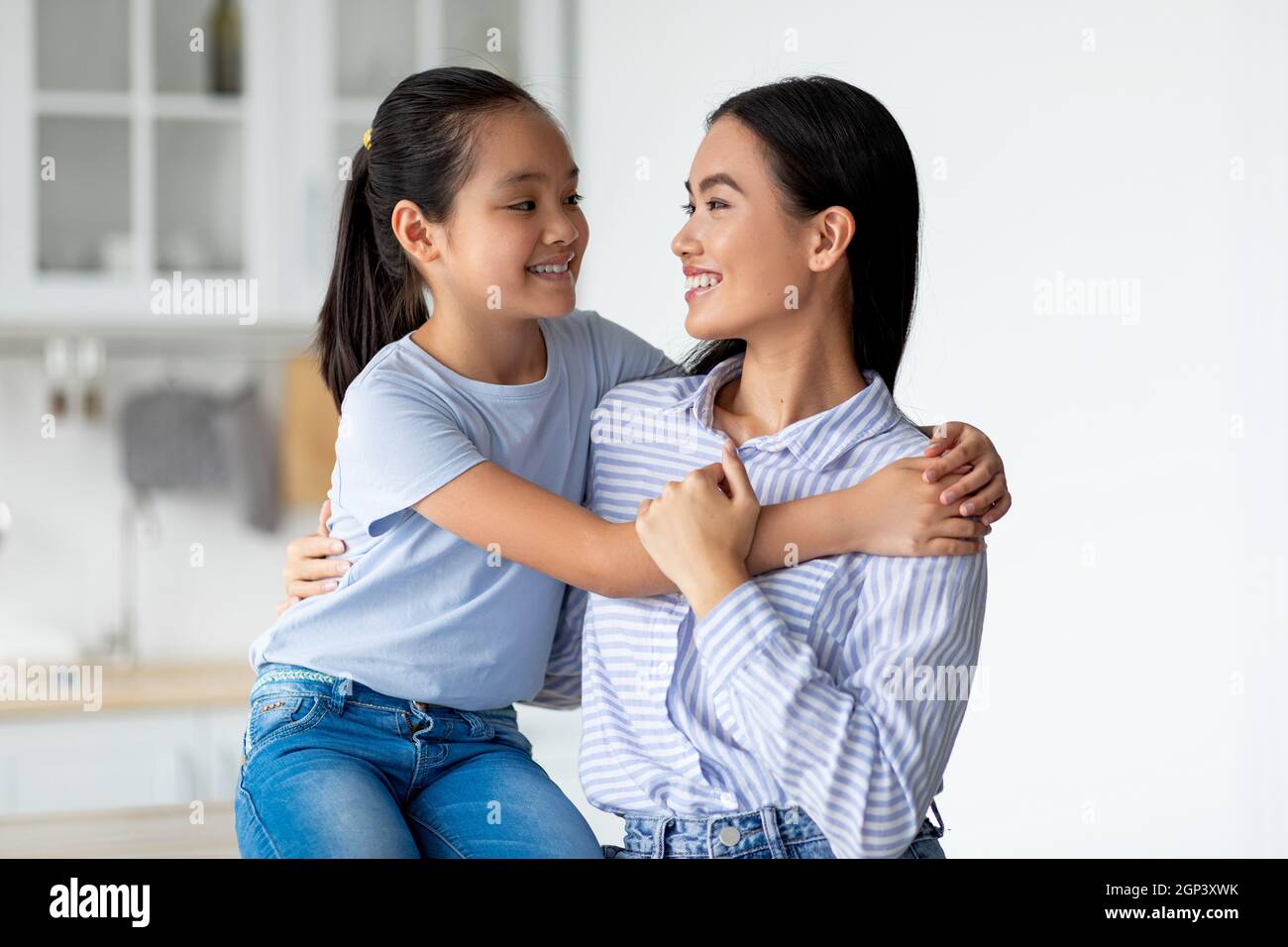 Mutter Tochter Liebe. Glückliche asiatische Dame, die sich mit ihrem Kind umarmt und in der Küche sitzt. Lächelndes Mädchen umarmt ihre Mutter zu Hause und begrüßt sie mit Motte Stockfoto