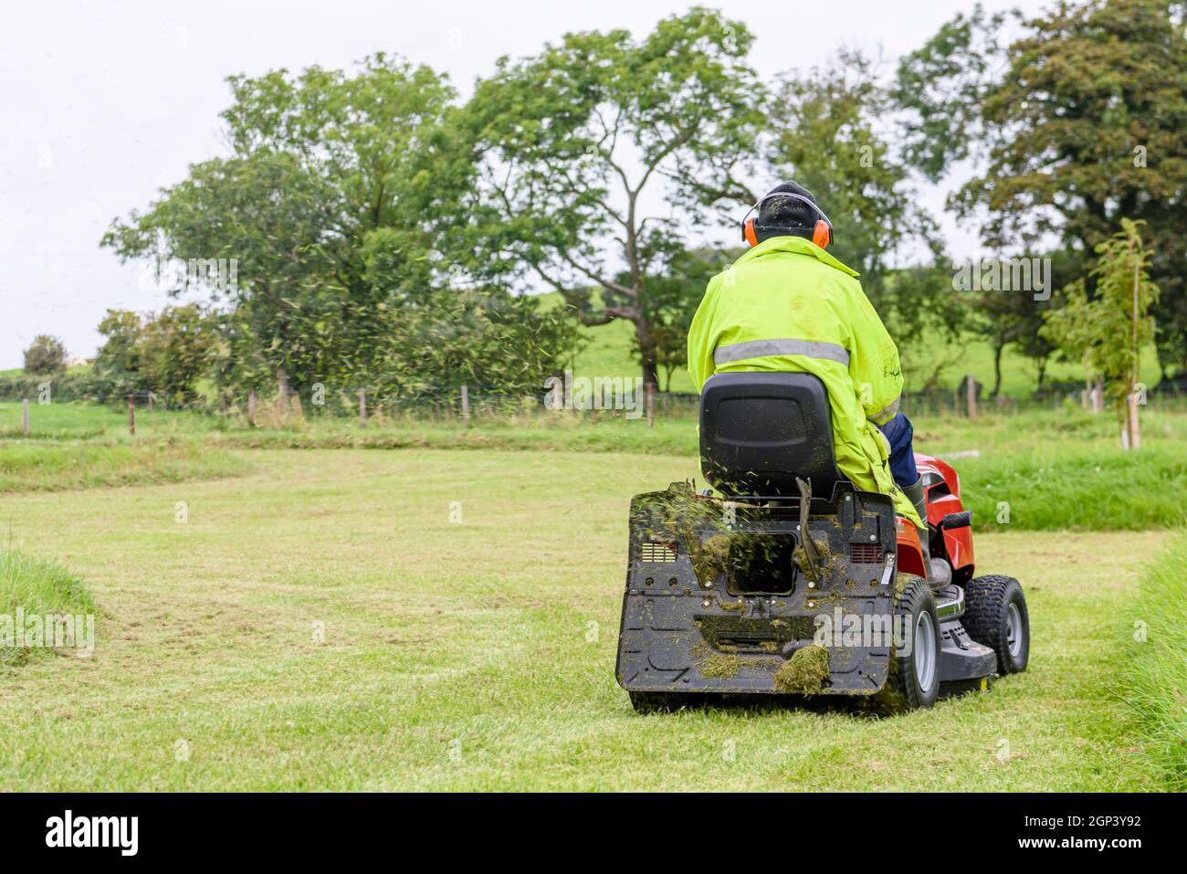 Ein Mann mit einer gut sichtbaren Jacke und Ohrenschützern mäht das Gras in einem großen Garten, während er auf einem Aufsitzmäher sitzt Stockfoto