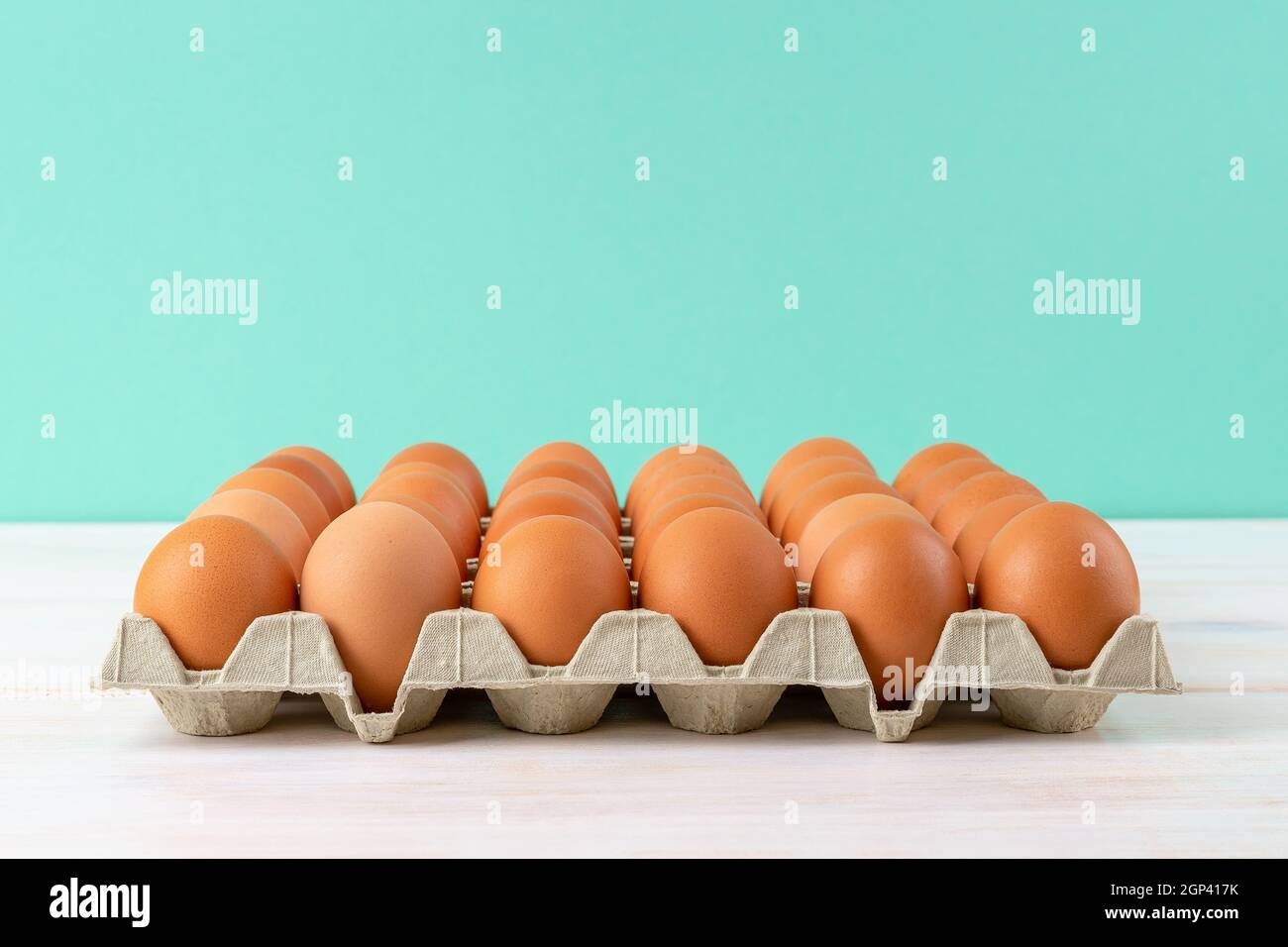 Braune Hühnereier im Papptablett auf einem weißen Holztisch vor türkisfarbenem Hintergrund. Rohe Bio-Hühnereier mit brauner Schale für Ostern. Stockfoto