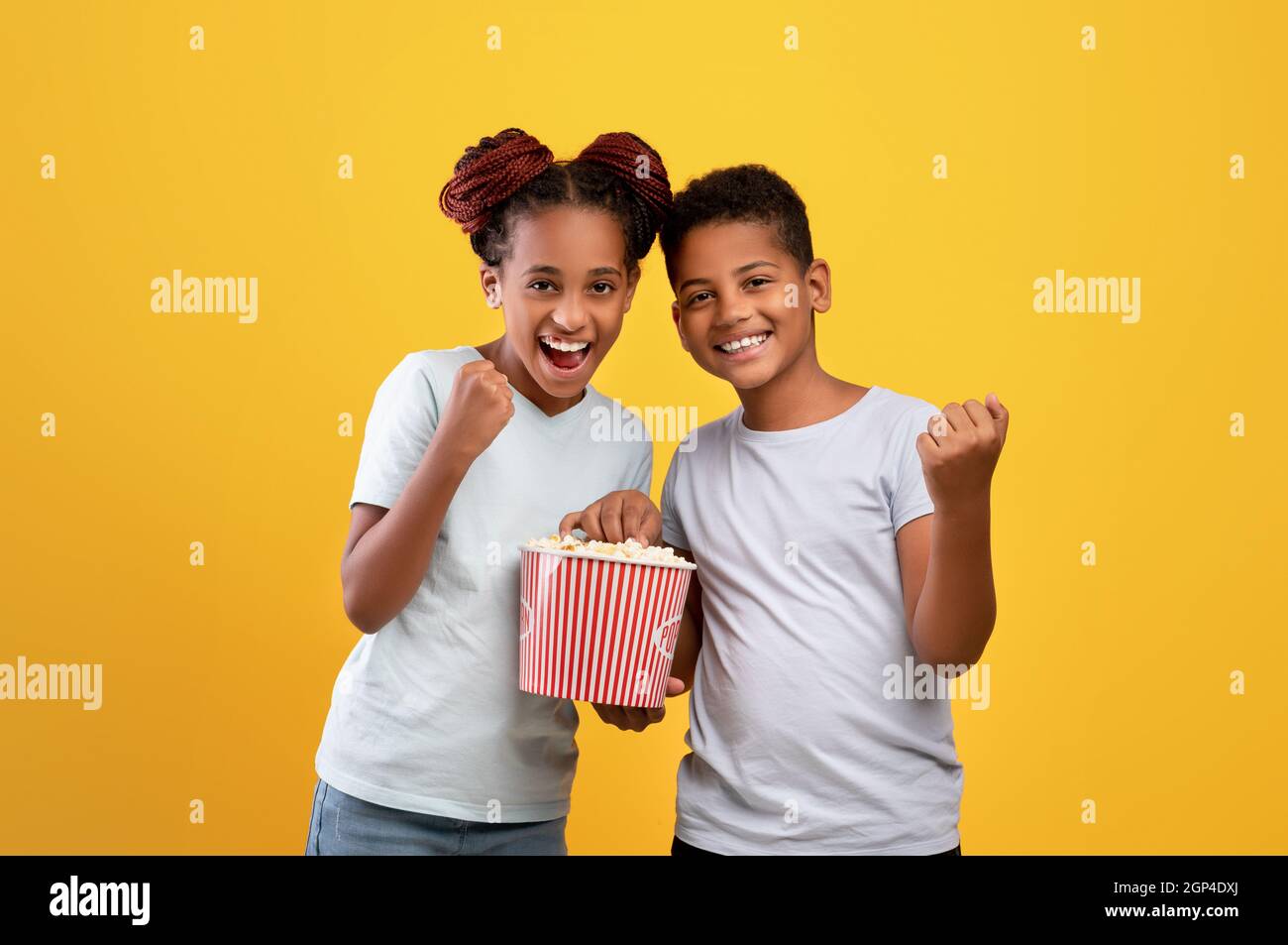 Fröhlicher schwarzer Bruder und Schwester, die zusammen im Kino Cartoon oder einen schönen Film sehen, Popcorn essen, geballte Fäuste hochheben, gelber Studiohintergrund Stockfoto