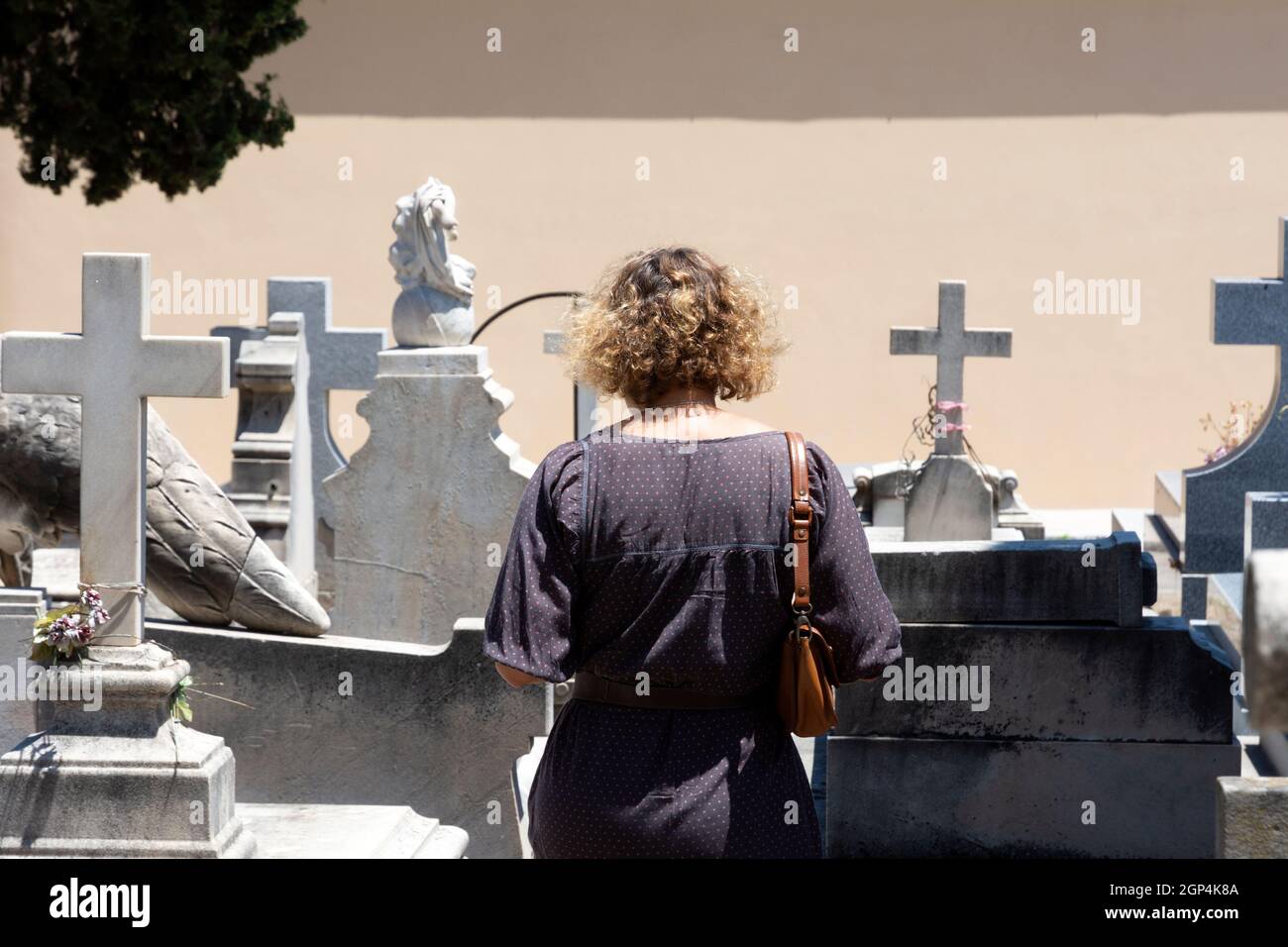 Nicht erkennbare, lockig blonde Frau, die ein Grab auf dem Friedhof besucht, mit mehreren Gräbern im Hintergrund und einer cremefarbenen Wand. Konzept von Stockfoto