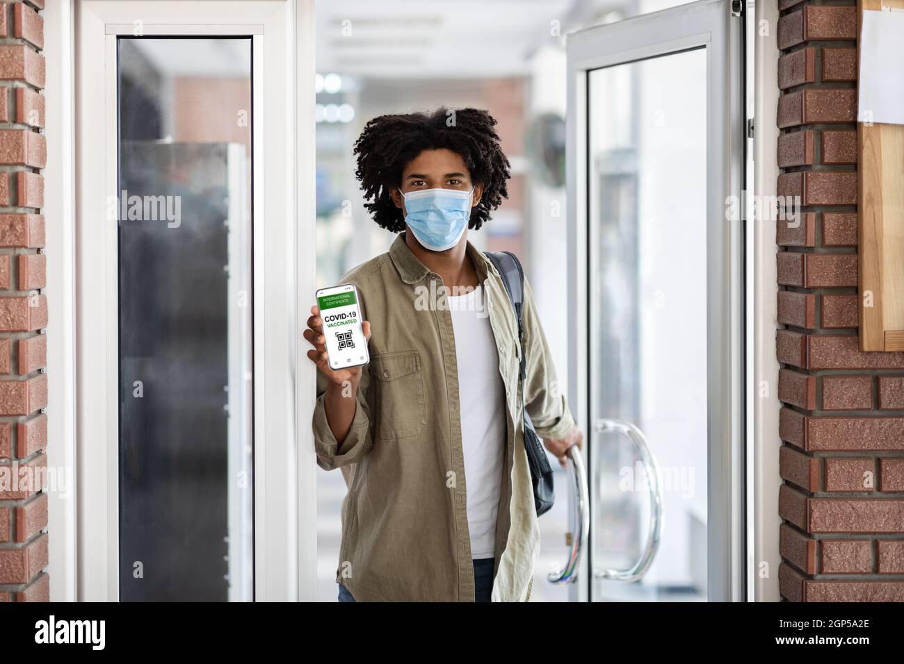 Schwarzer Mann In Schützender Medizinischer Maske Zeigt Digitales Impfzertifikat Beim Betreten Des Gebäudes, Junger Afroamerikanischer Mann Demonstriert Smartph Stockfoto