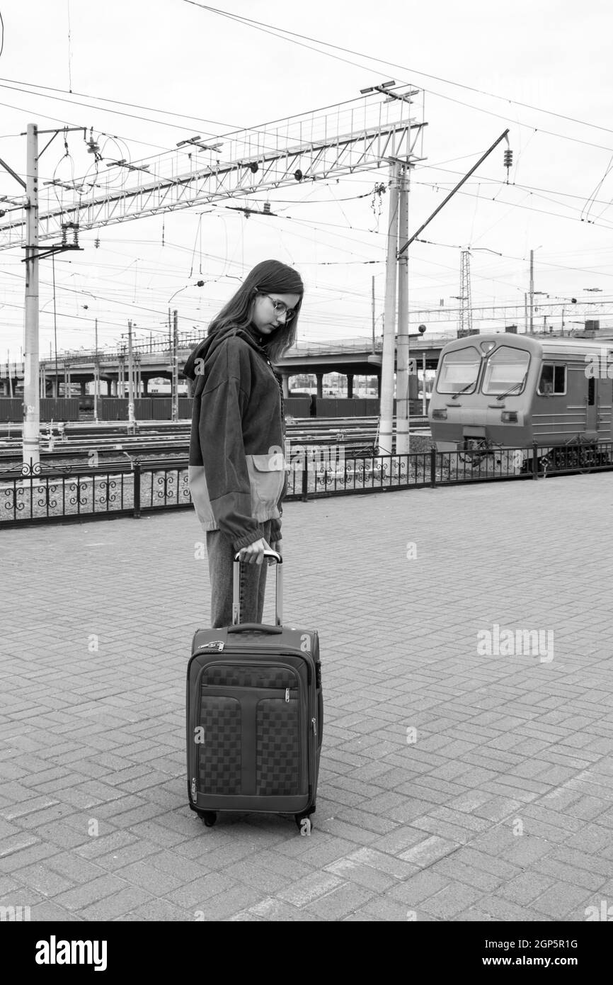 Kaukasische Schülerin in heller Kleidung mit einem Koffer, der zum Elektrozug am Bahnhof geht, schwarz und weiß. Mädchen mit einem Koffer am Trai Stockfoto