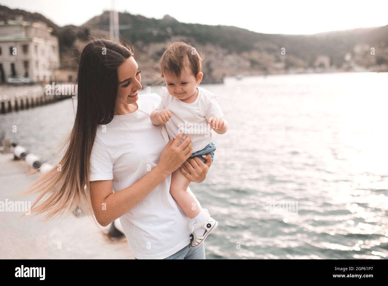 Junge Mutter halten Baby Mädchen 1 Jahr alt tragen legere Kleidung Spaziergang in der Stadt Straße über das Meer. Mutterschaft. Glückliche Frau mit Kind zusammen draußen. Stockfoto