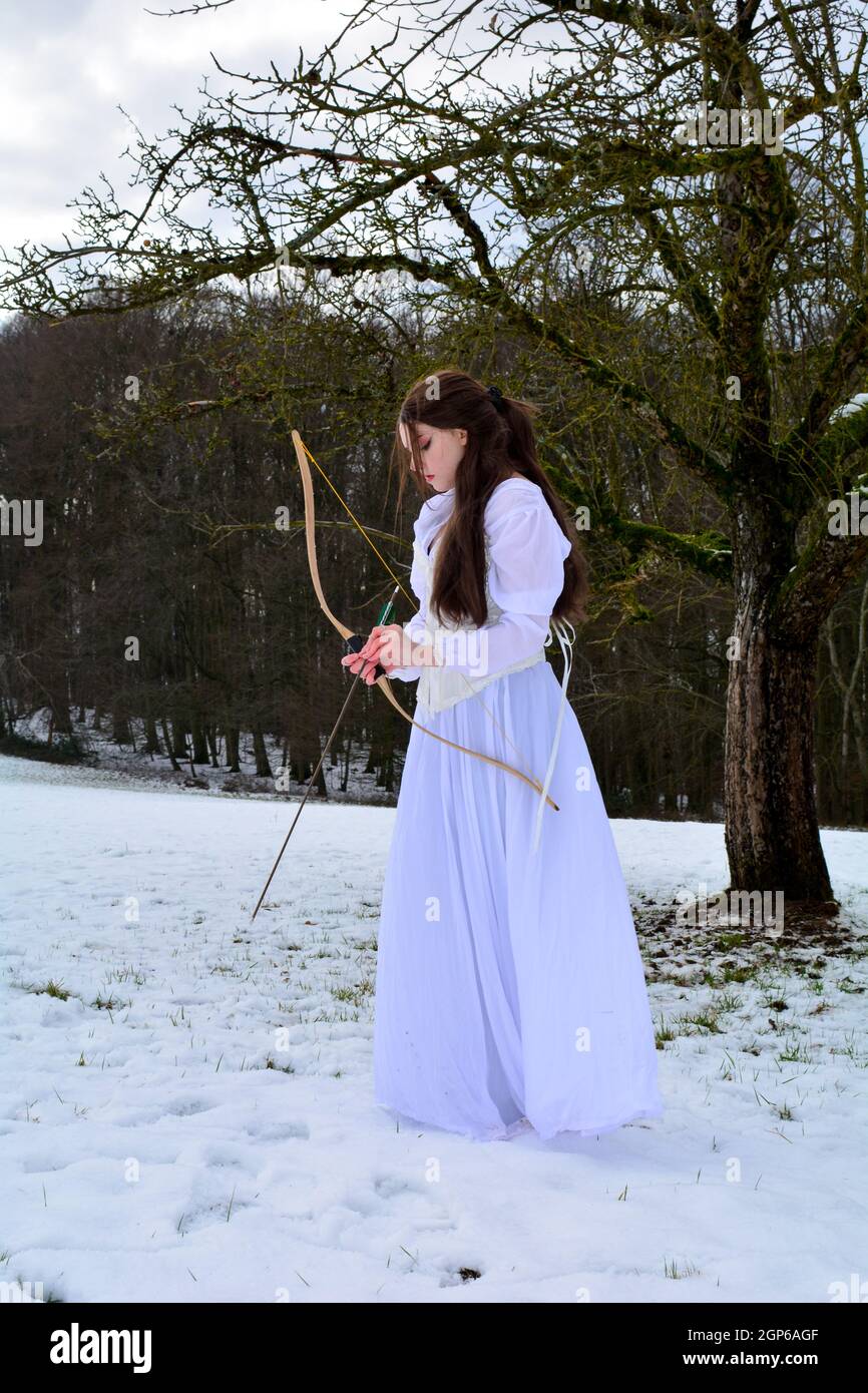 Junge Frau steht seitlich mit langem weißen Kleid im Schnee vor einem Wald, spielt mit Pfeil und Bogen Stockfoto