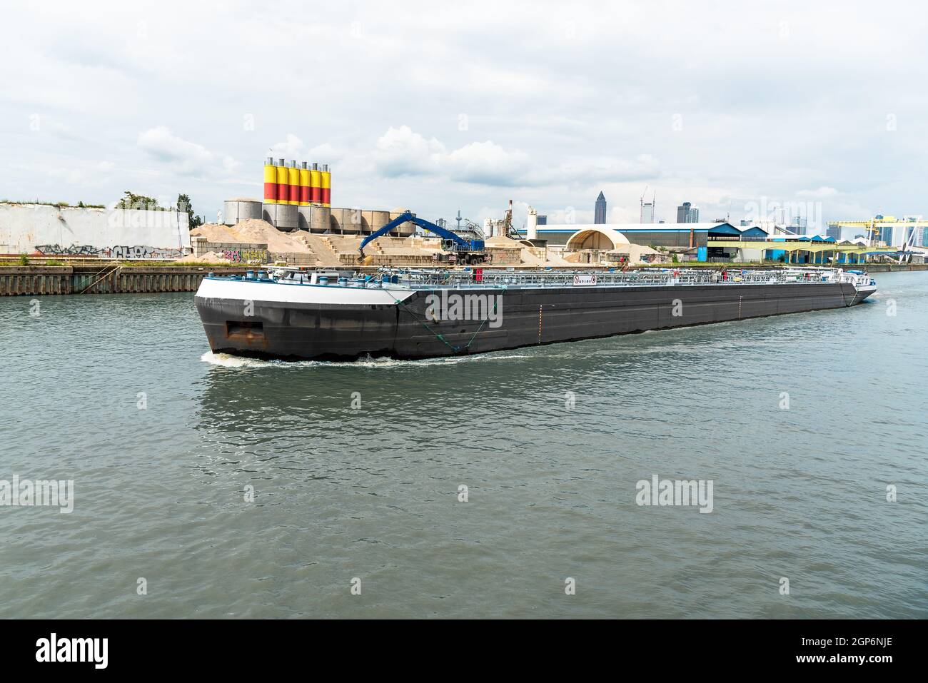 An einem bewölkten Sommertag segelt ein Tankerschiff auf einem Fluss durch ein Industriegebiet Stockfoto