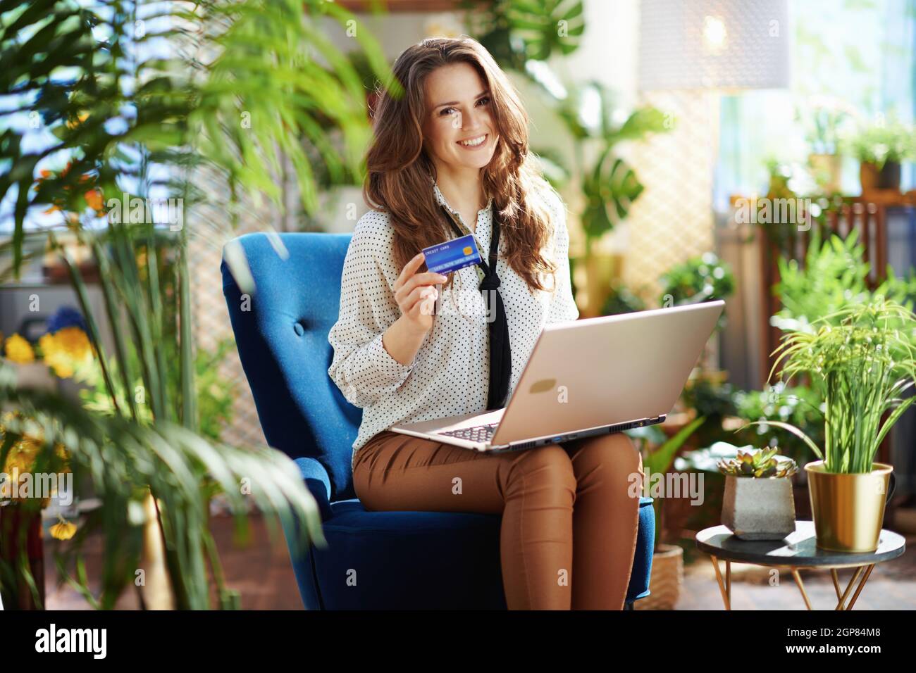 Green Home. Lächelnde junge Frau mit langen welligen Haaren mit Laptop und Kreditkarte in beigen Hosen und Bluse sitzt in einem blauen Sessel in der modernen Hou Stockfoto