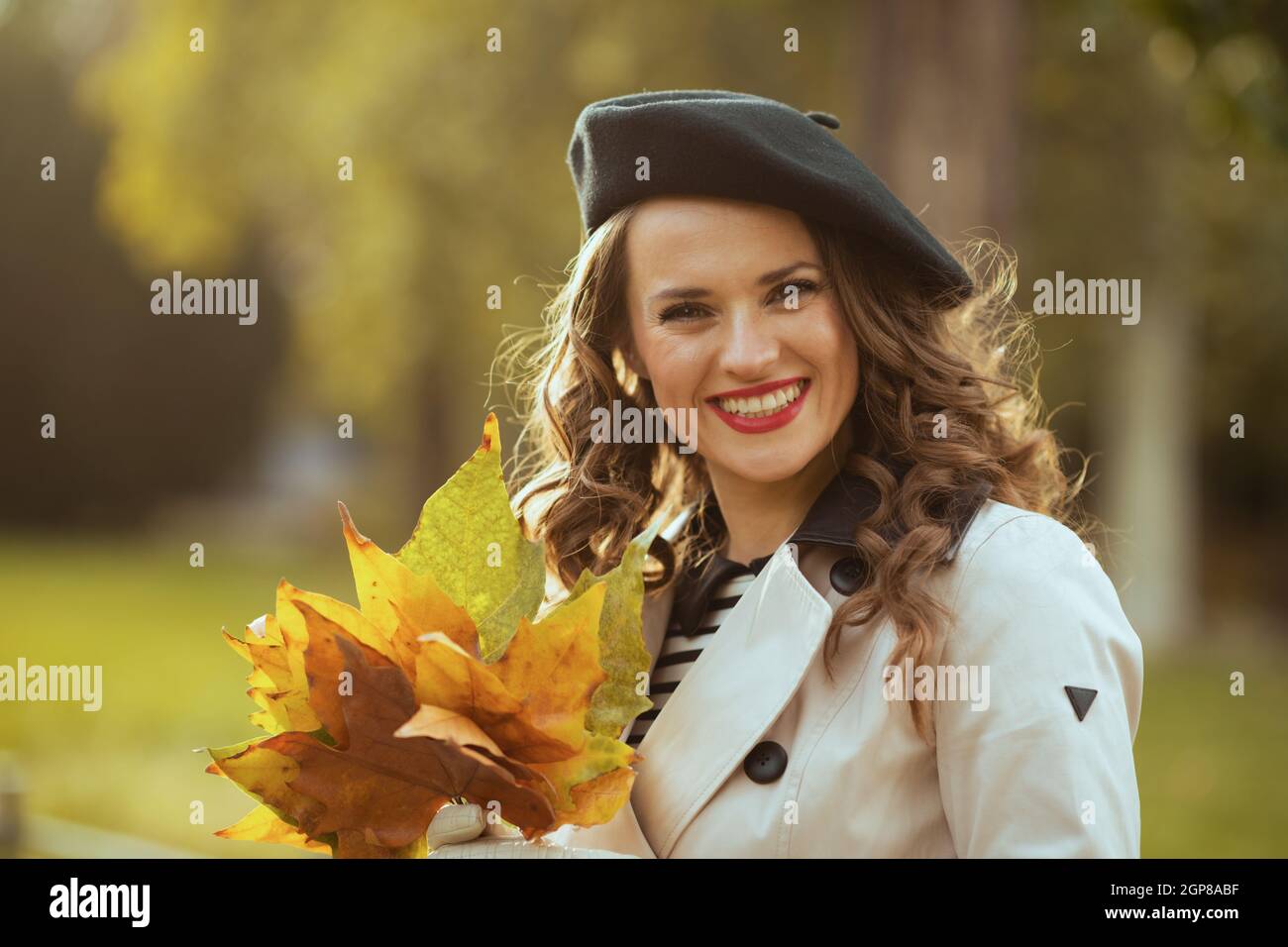 Hallo oktober. Porträt einer glücklichen trendigen Frau im beigen Trenchcoat mit herbstlichen gelben Blättern draußen in der Stadt im Herbst. Stockfoto