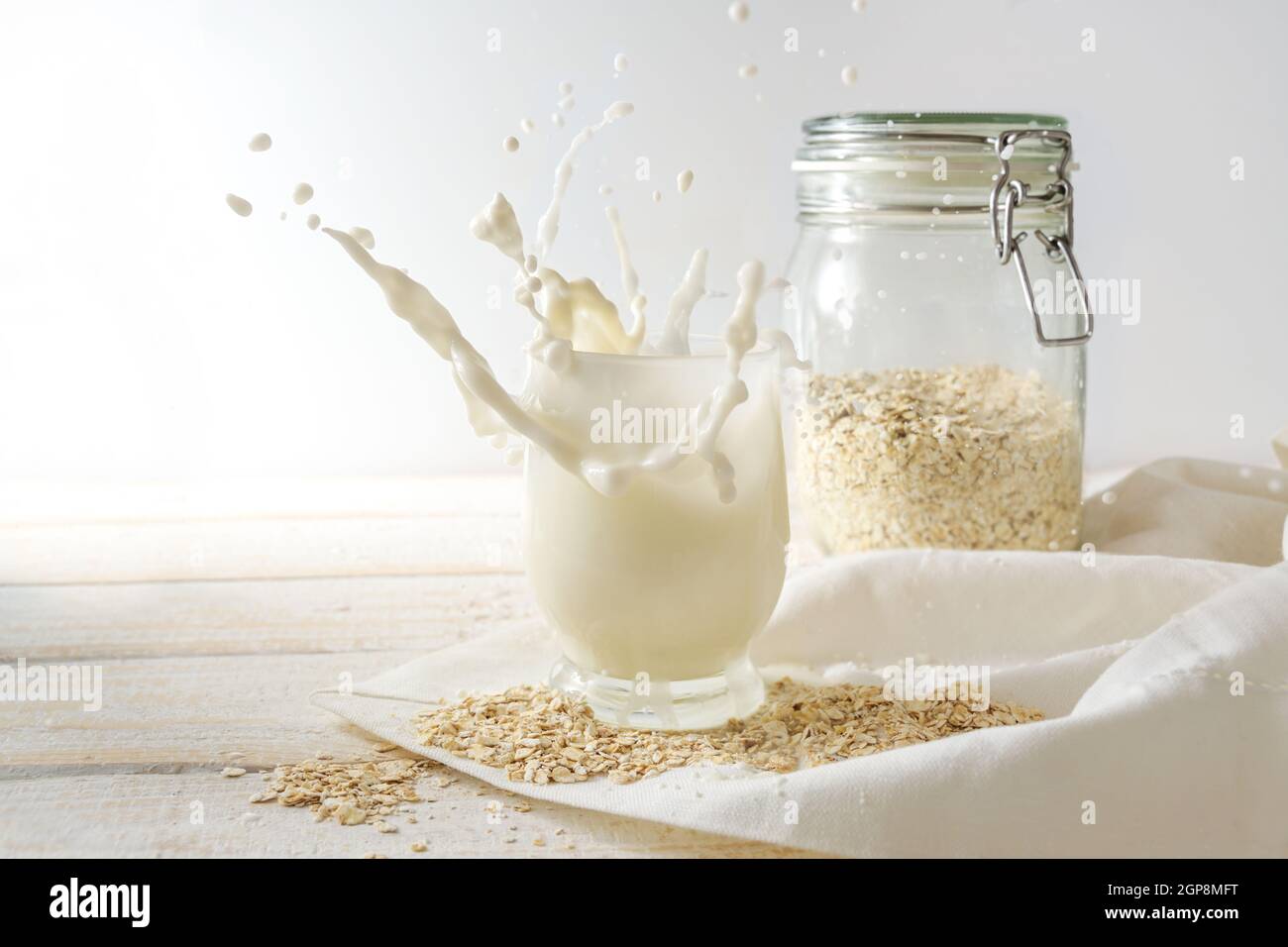 Vegane Hafermilch spritzt in ein Trinkglas und ein Vorratsglas auf einem hellen Holztisch mit weißer Serviette, gesund laktosefrei und alternativ nicht milchfrei Stockfoto